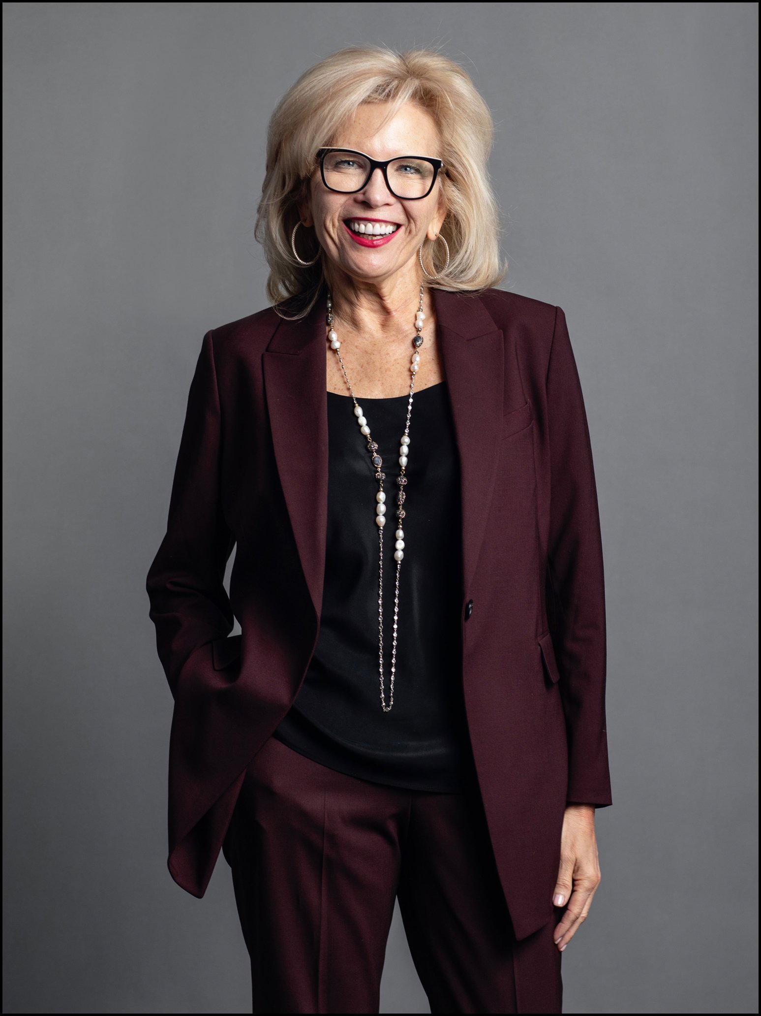 Smiling woman with blonde hair, wearing glasses, a burgundy suit, black top, and jewelry, standing against a gray background.