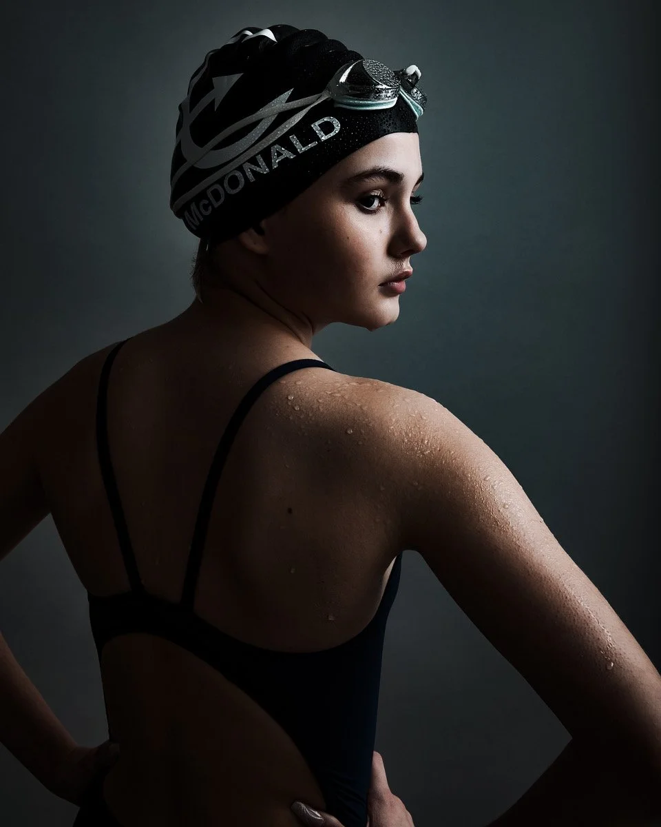 A young female swimmer wearing a black swim cap with goggles on top, in a dark and moody setting, with water droplets on her shoulder and arm, looking to the side.