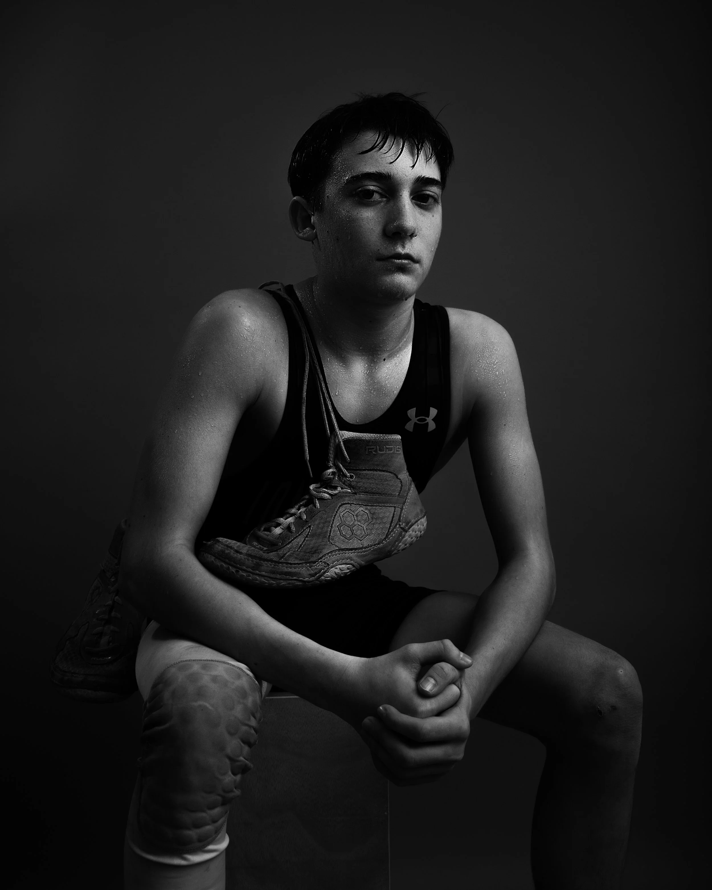 Black and white photo of a young male athlete sitting on a stool, wearing athletic gear, with a serious expression, and sweat visible on his face and arms.