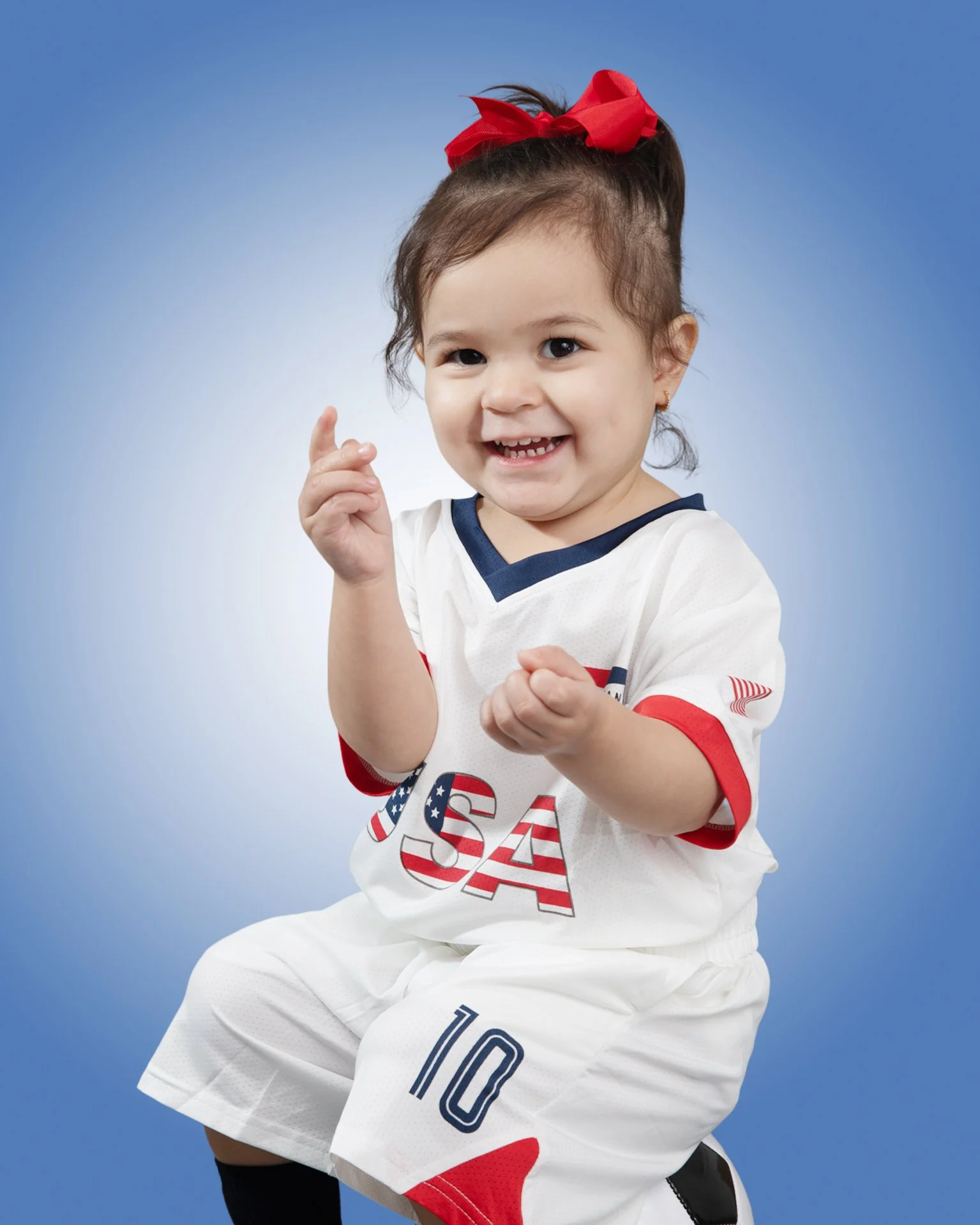 Smiling young girl with brown hair in a ponytail with a red bow, wearing a white sports uniform with USA lettering and number 10, sitting on a black stool against a blue gradient background.