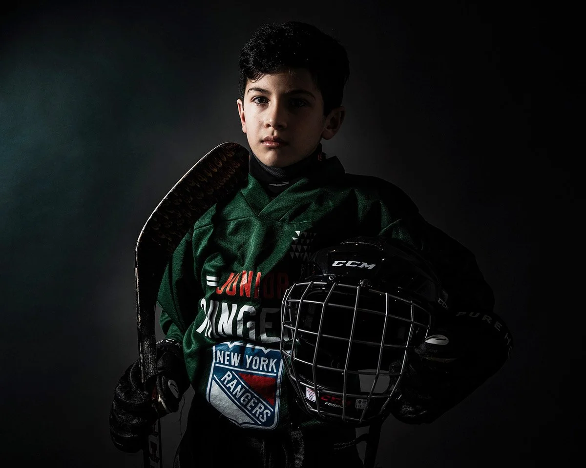 Young boy in hockey gear holding a hockey stick and helmet against a dark background.