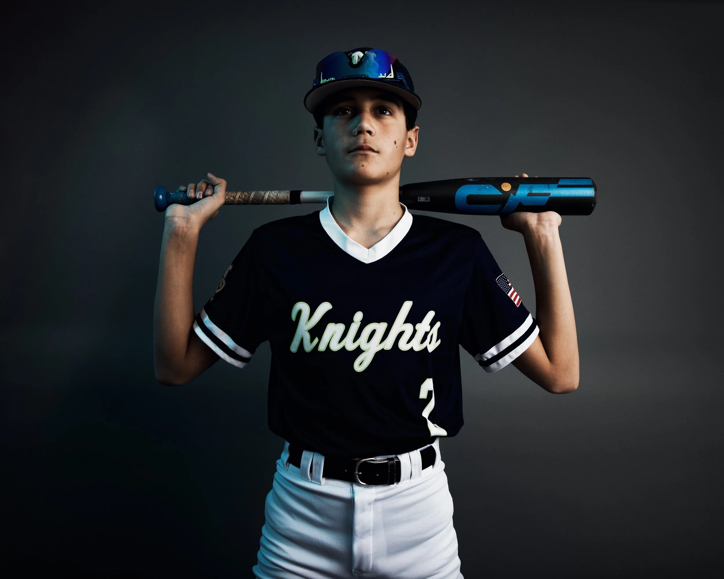A teenage boy wearing a black baseball jersey with 'Knights' written on it, white baseball pants, a black belt, a black cap with sunglasses on top, and holding a bat over his shoulders.