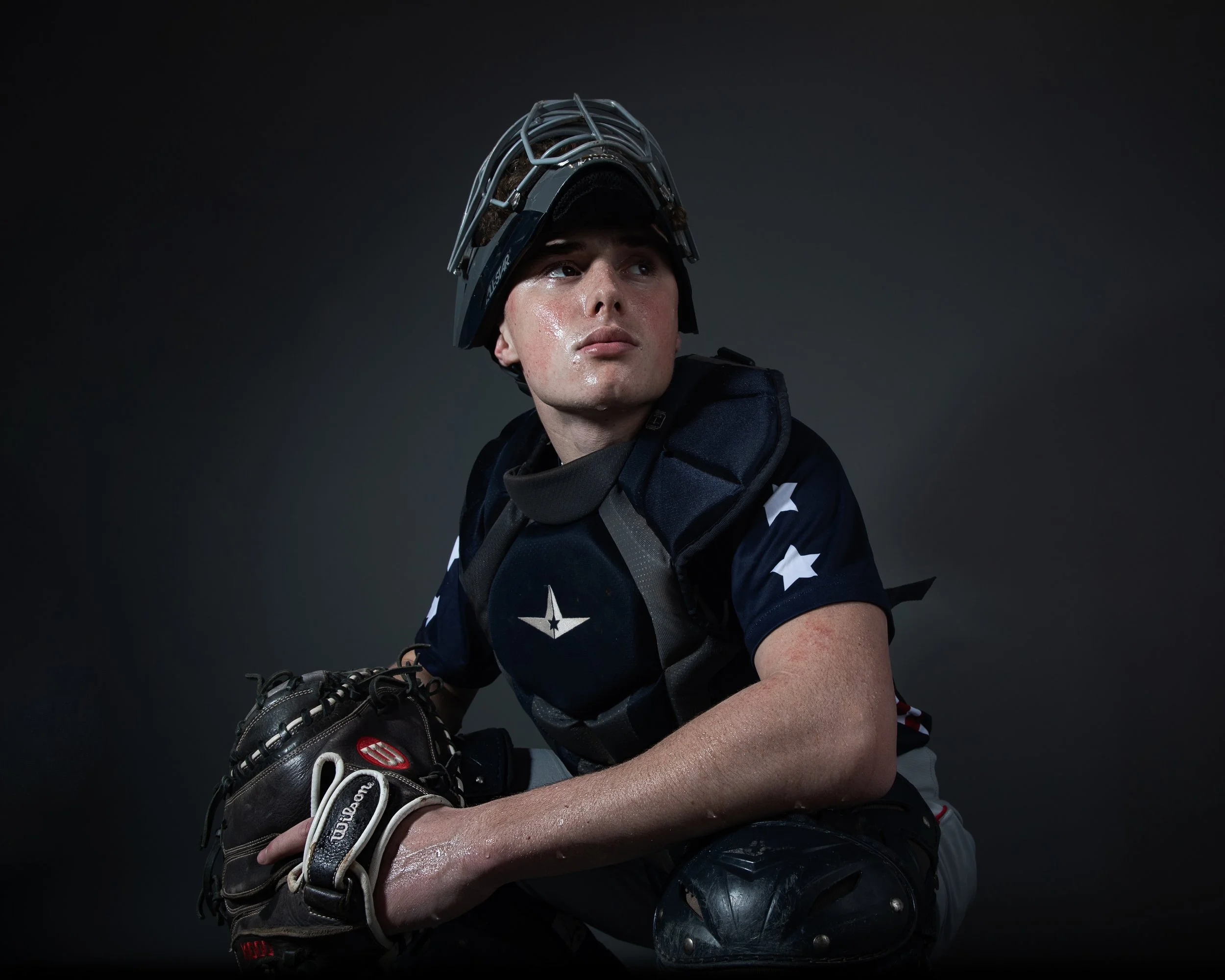 A young male baseball player dressed in uniform with stars, wearing a catcher's helmet, kneeling with a glove and protective gear.