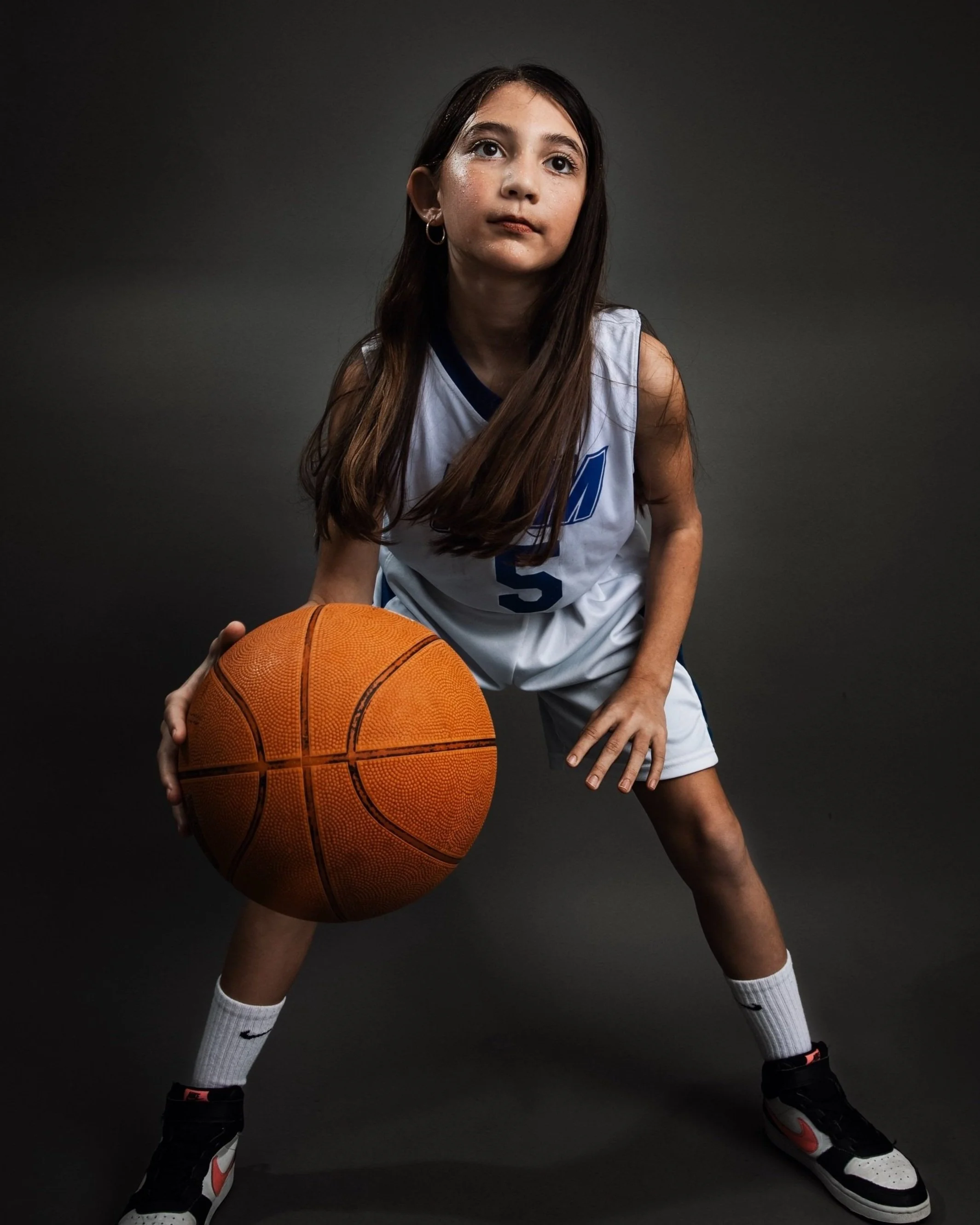 A young girl in a basketball jersey holding a basketball, standing in a defensive stance against a dark background.
