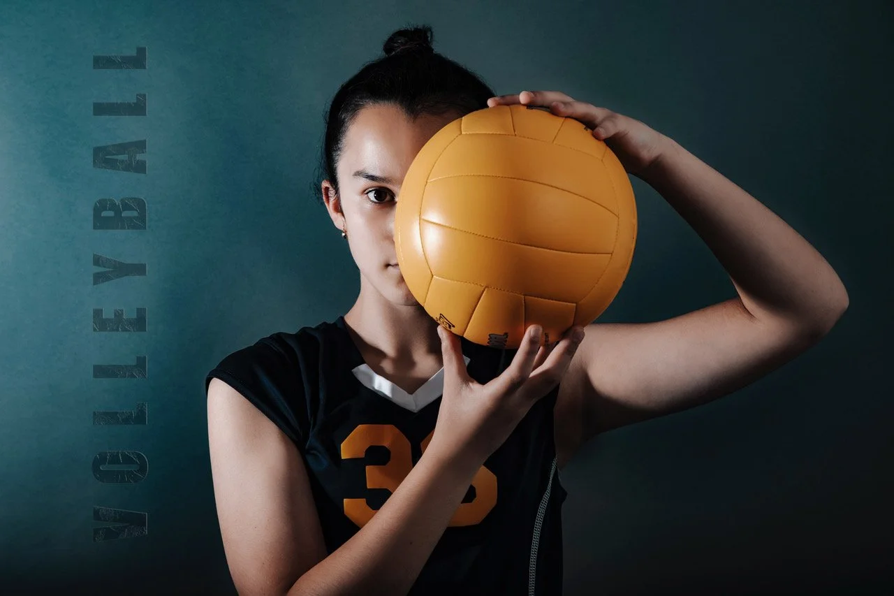 A woman in a sports jersey holding a yellow volleyball in front of her face against a dark teal background, with the word 'VOLLEYBALL' written vertically on the left side.