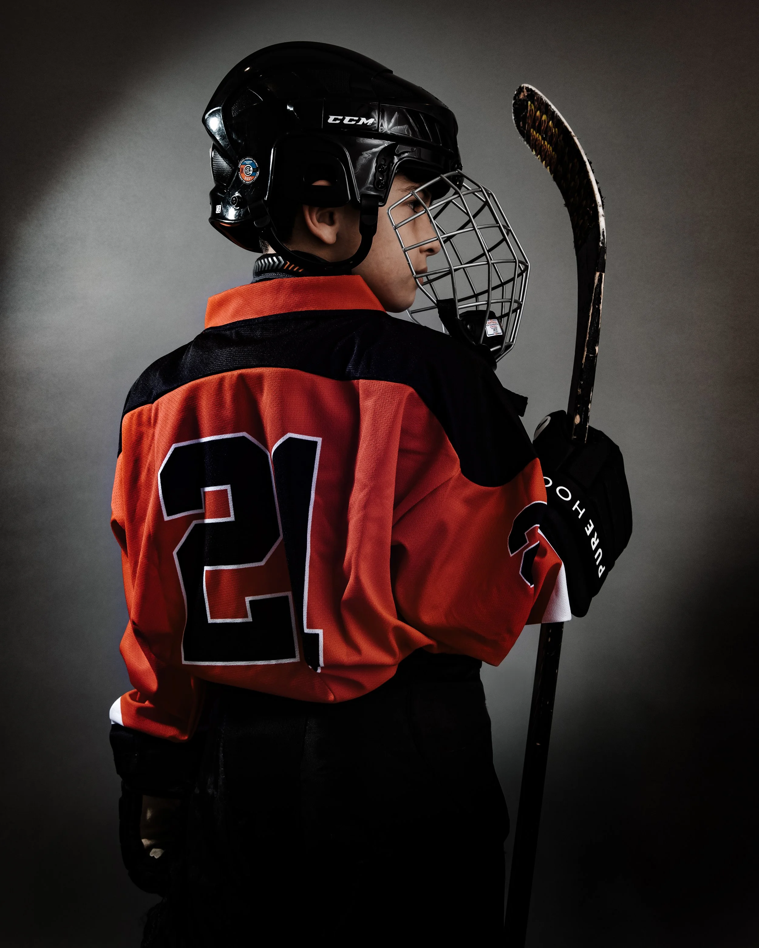 A young hockey player in a red and black jersey with the number 21, wearing a black helmet and holding a hockey stick, facing sideways.