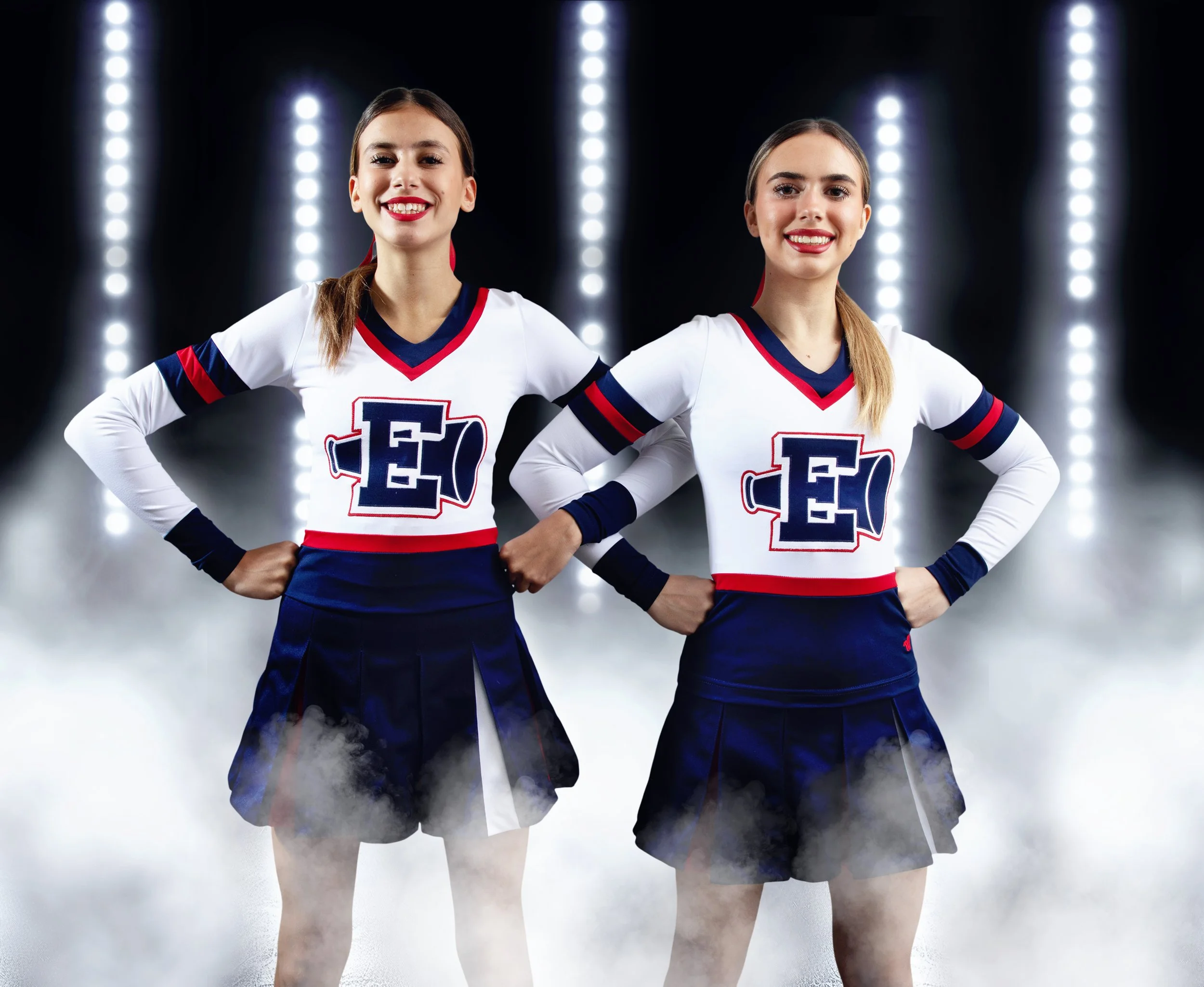 Two young female cheerleaders in matching white, navy, and red uniforms with a large letter 'E' on the front, standing with hands on hips in a smoky, well-lit stadium or performance setting.