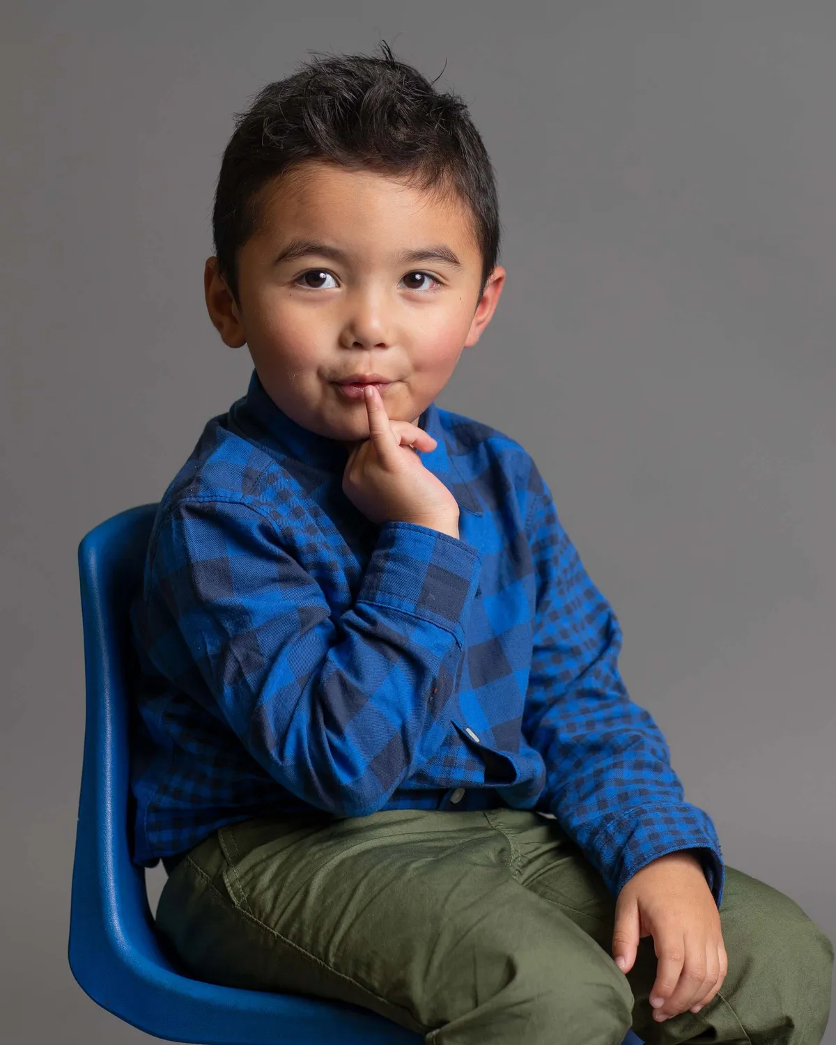 A young boy sitting on a blue chair with a gray background, wearing a blue plaid shirt and green pants, holding his finger to his lips in a thoughtful pose.