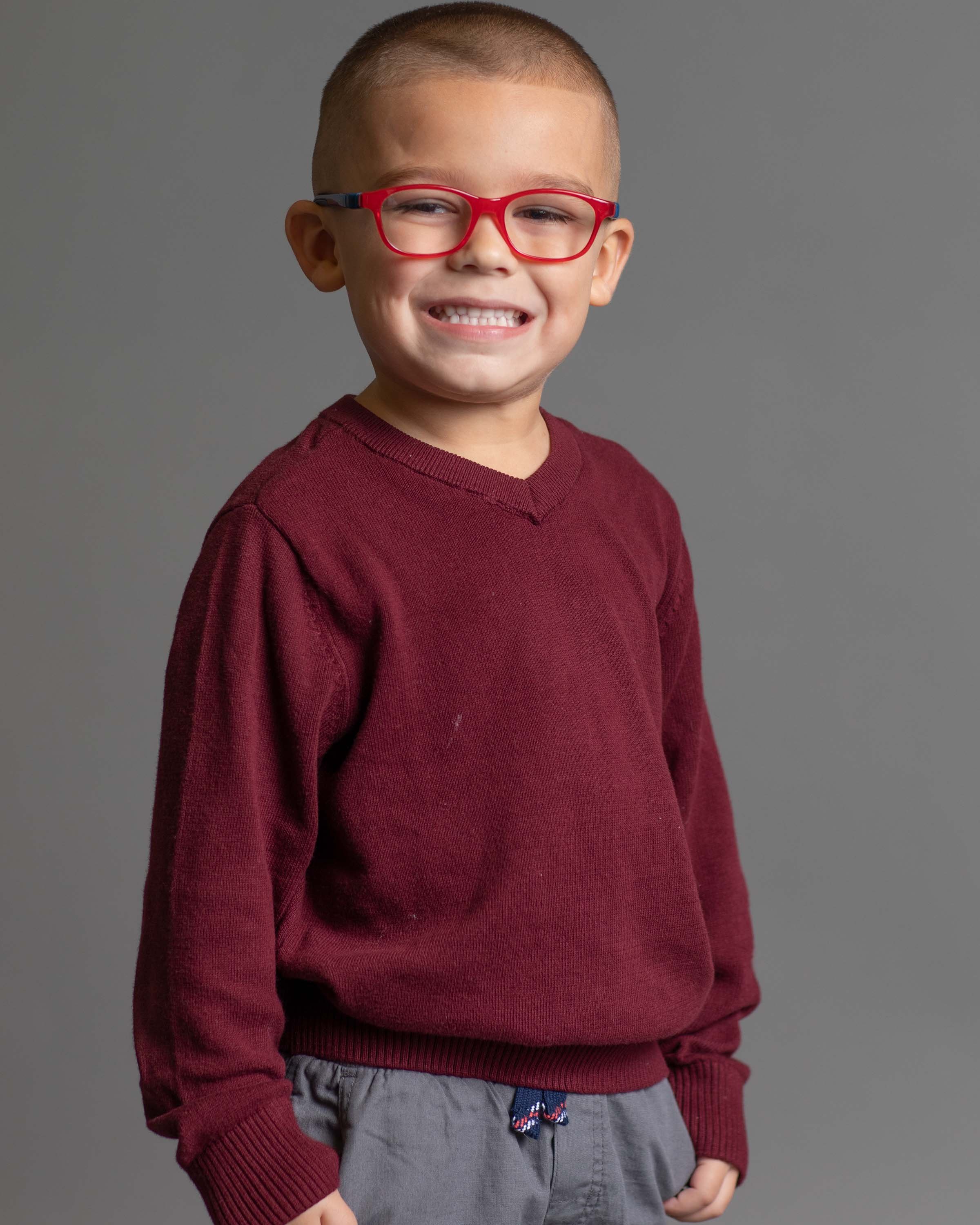 A young boy smiling, wearing red glasses, a maroon sweater, and gray pants against a gray background.
