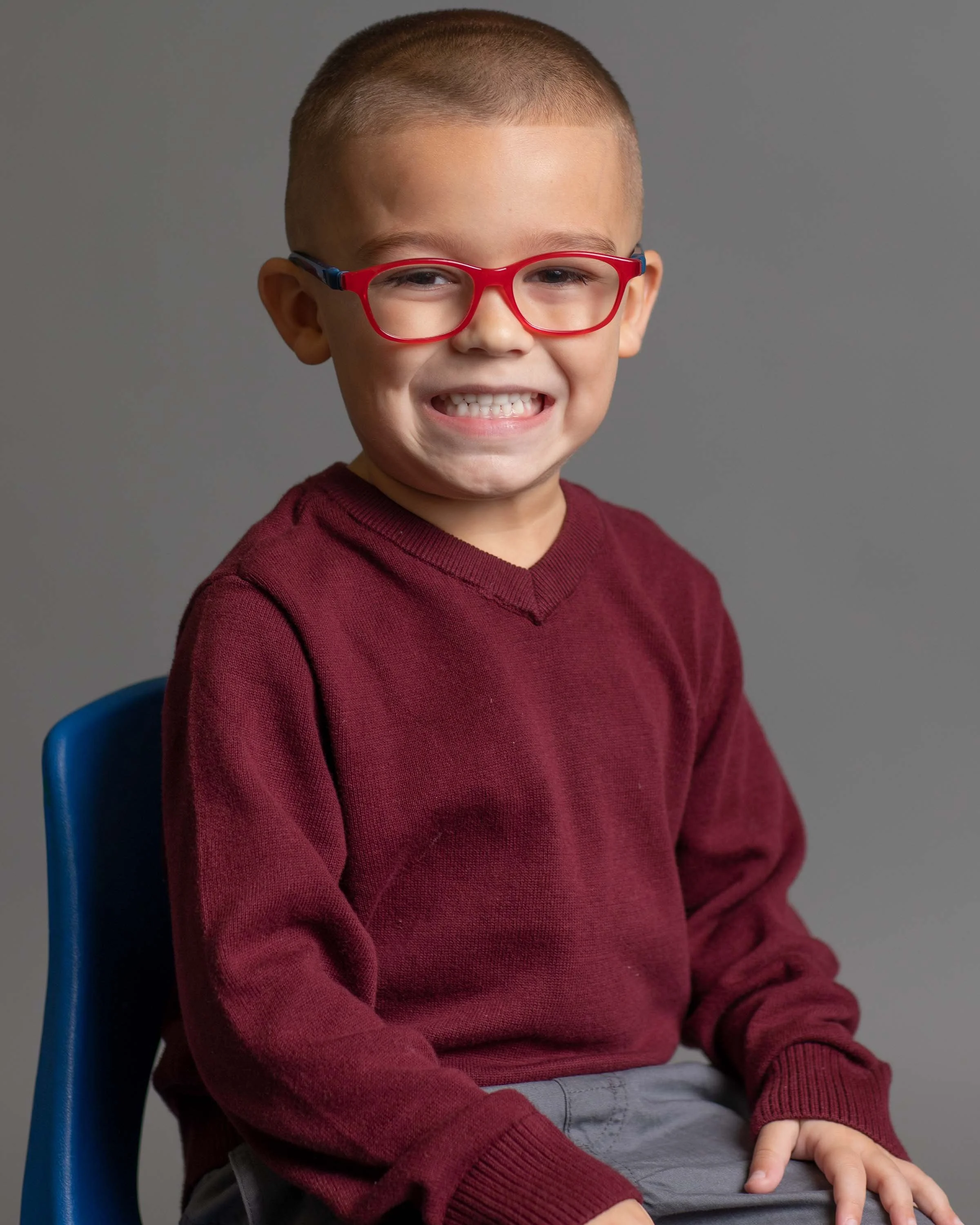 A young boy wearing red glasses, a maroon sweater, and gray pants, sitting on a blue chair, smiling at the camera against a gray background.