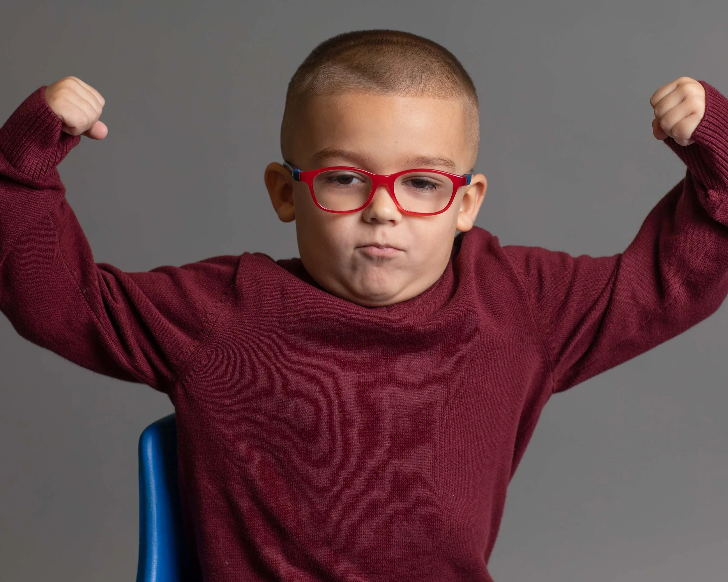 A young boy with short hair, wearing red glasses and a maroon sweater, flexing his arms and making a determined face against a plain gray background.