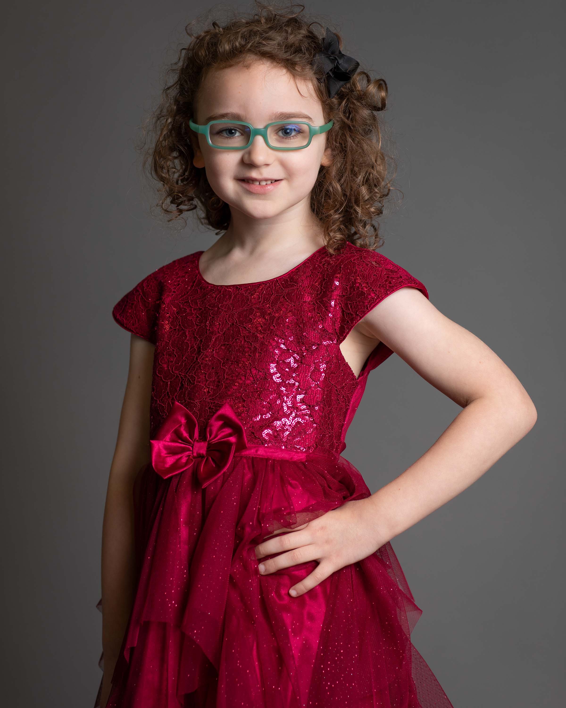 Young girl with curly hair, wearing glasses and a red, sparkly dress with a bow, smiling at the camera.