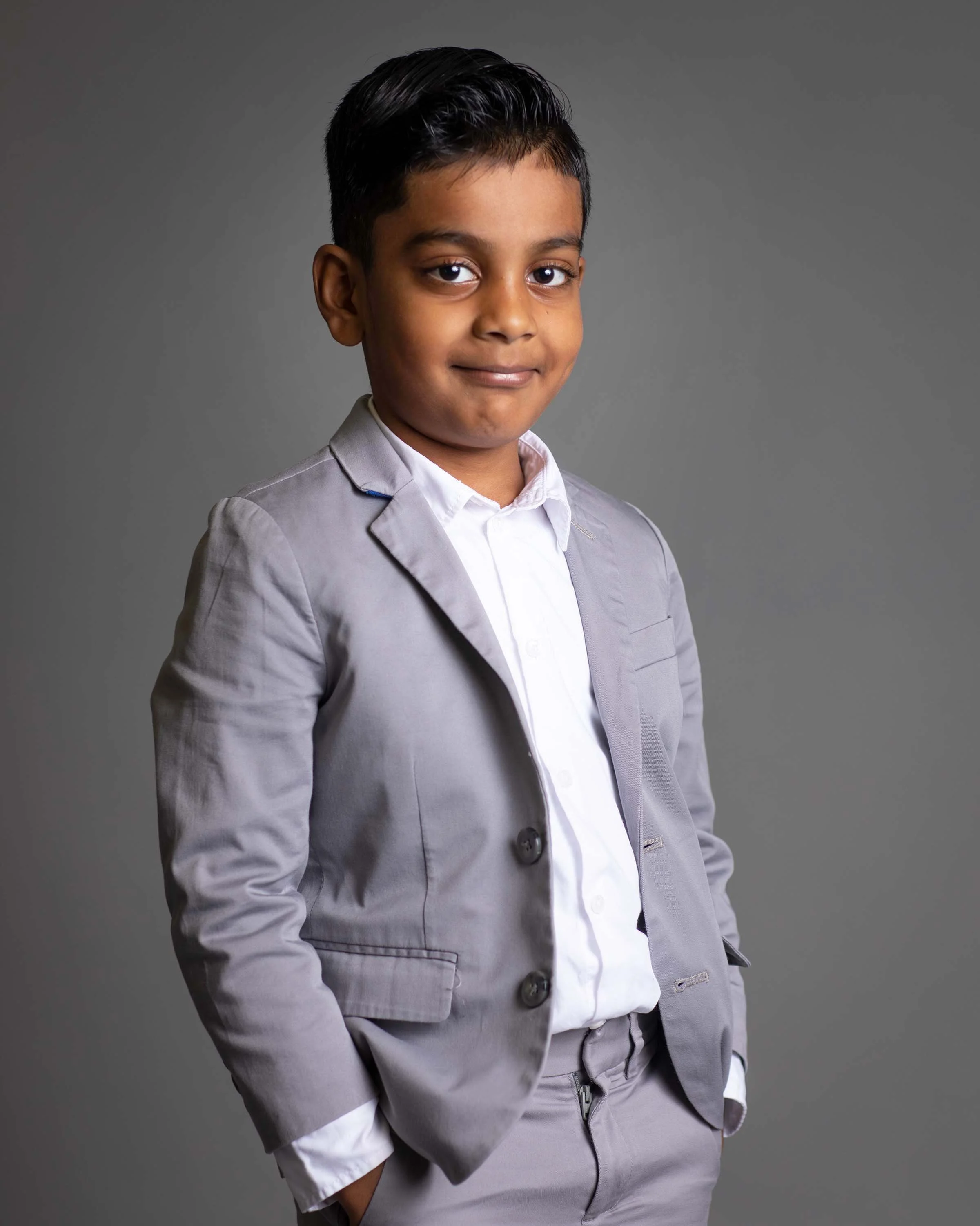 A young boy in a gray suit with a white shirt standing against a gray background.