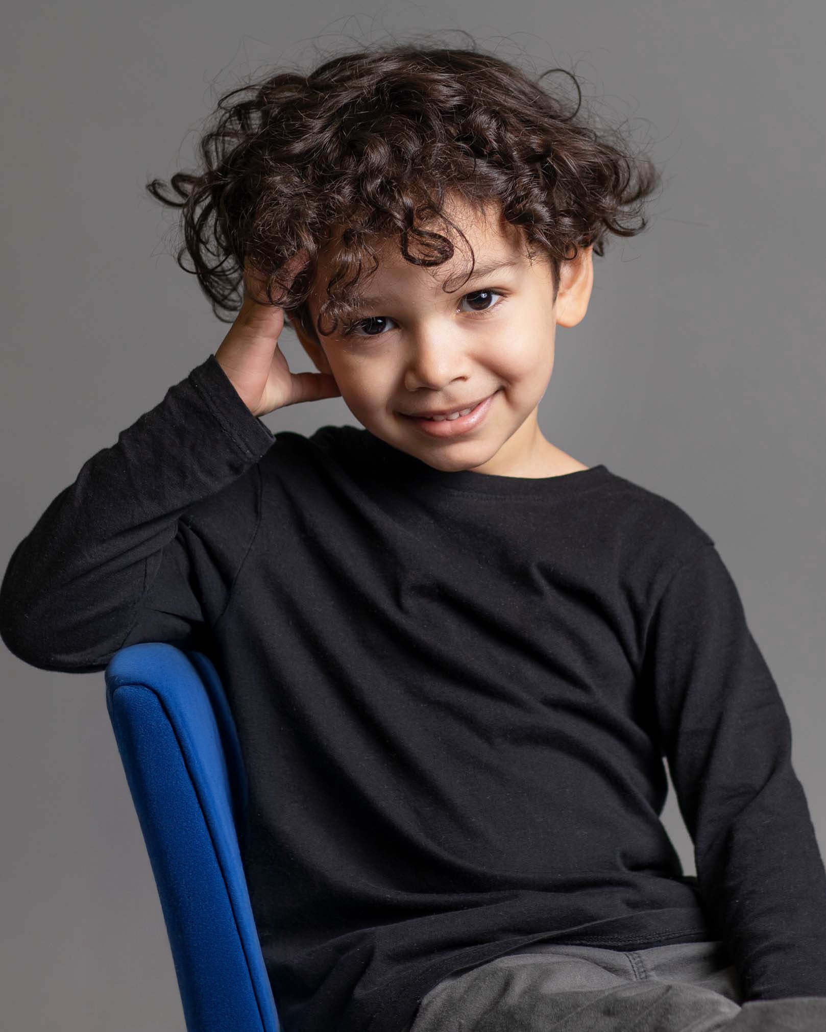 A young boy with curly brown hair and dark eyes, smiling and touching his head, sitting on a blue chair against a gray background.