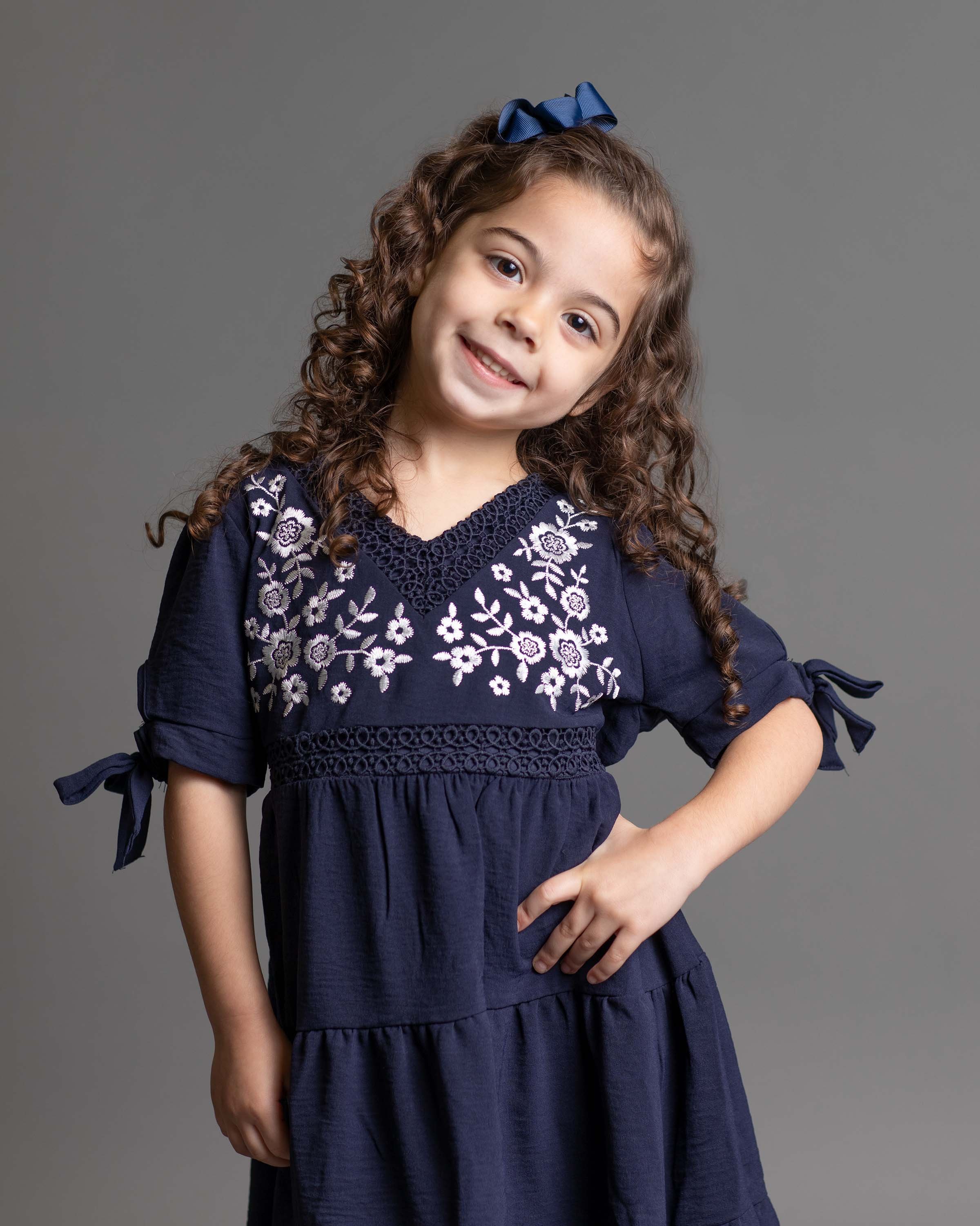 Young girl with curly brown hair, blue bow, in a navy dress with white embroidery, posing with one hand on her hip against a gray background.