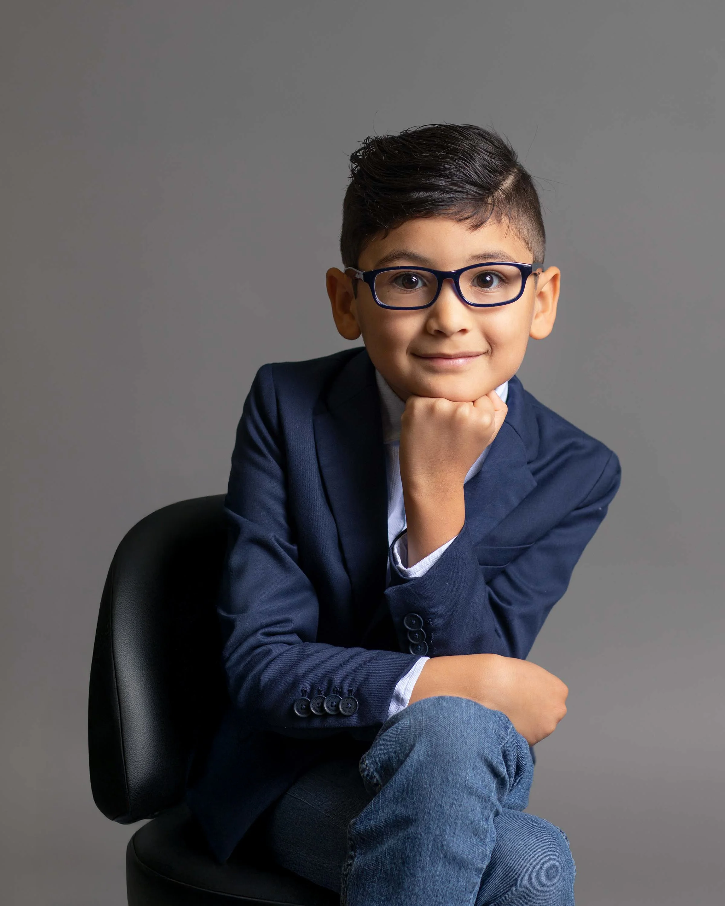 A young boy wearing glasses, a navy blazer, and jeans, sitting on a black chair against a gray background, resting his chin on his hand and smiling.