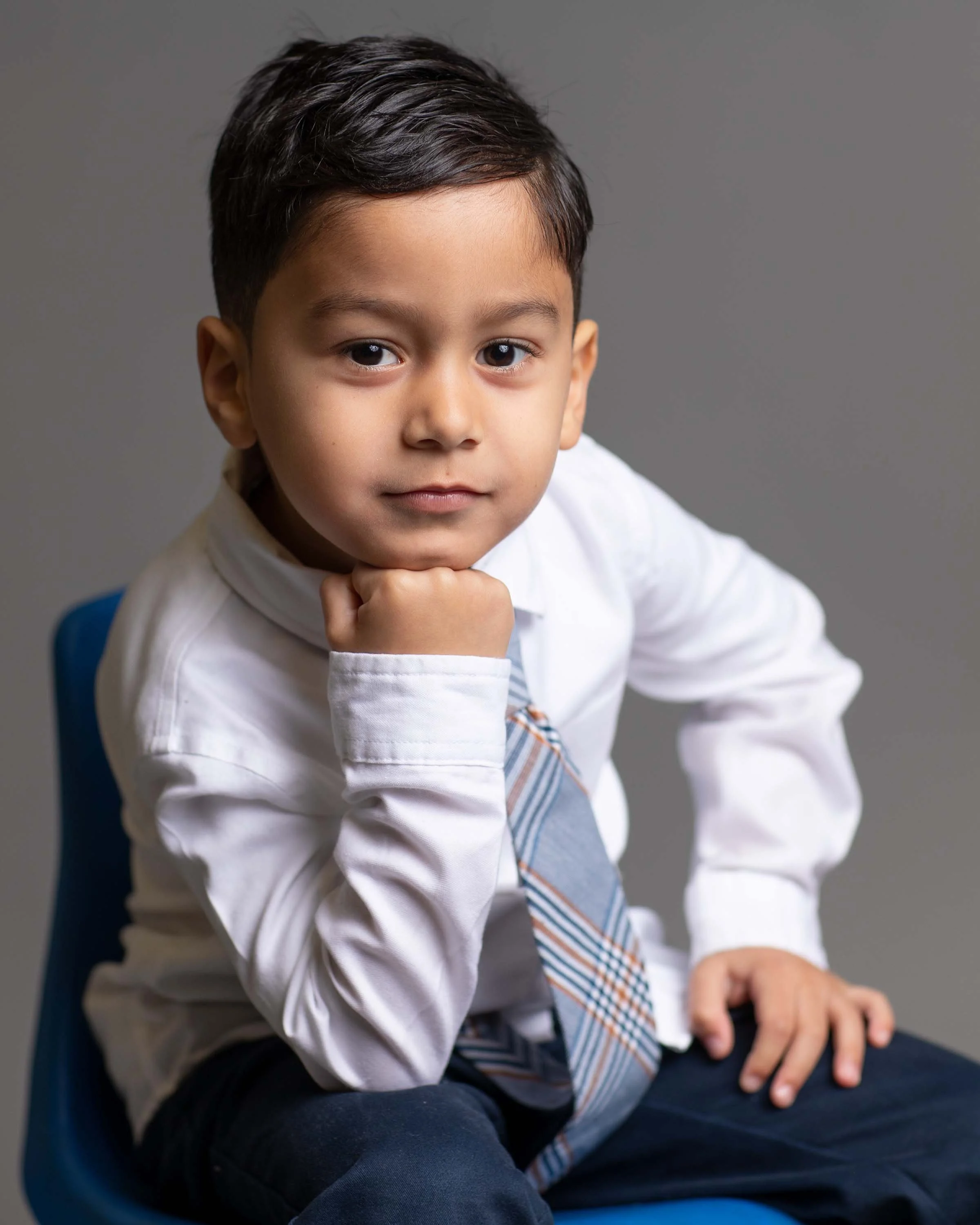 A young boy in a white shirt and striped tie sitting on a blue chair, resting his chin on his hand, looking confidently at the camera.