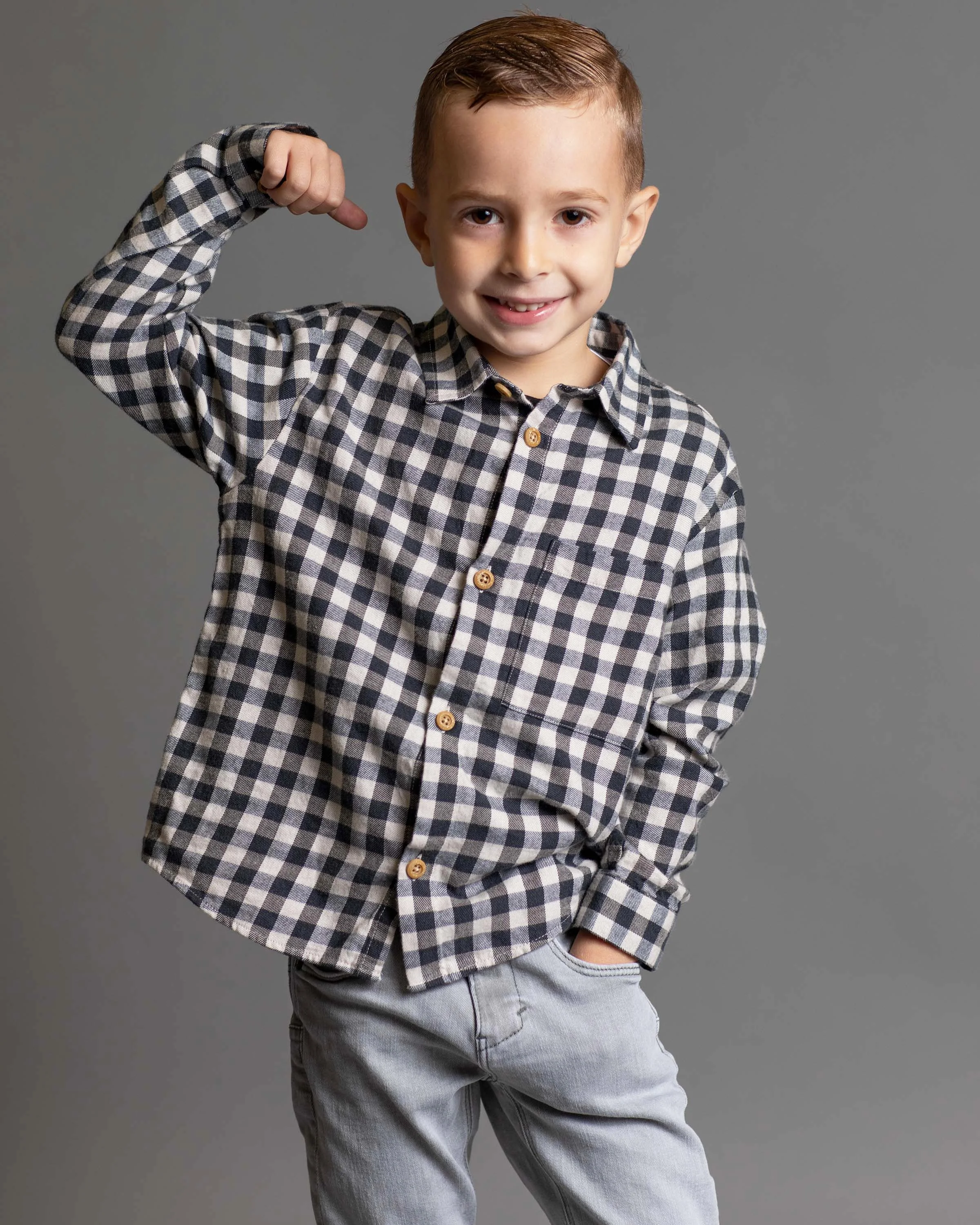 Young boy with short light brown hair smiling, wearing a black and white checkered shirt and light gray jeans, flexing his right arm in a pose against a plain gray background.
