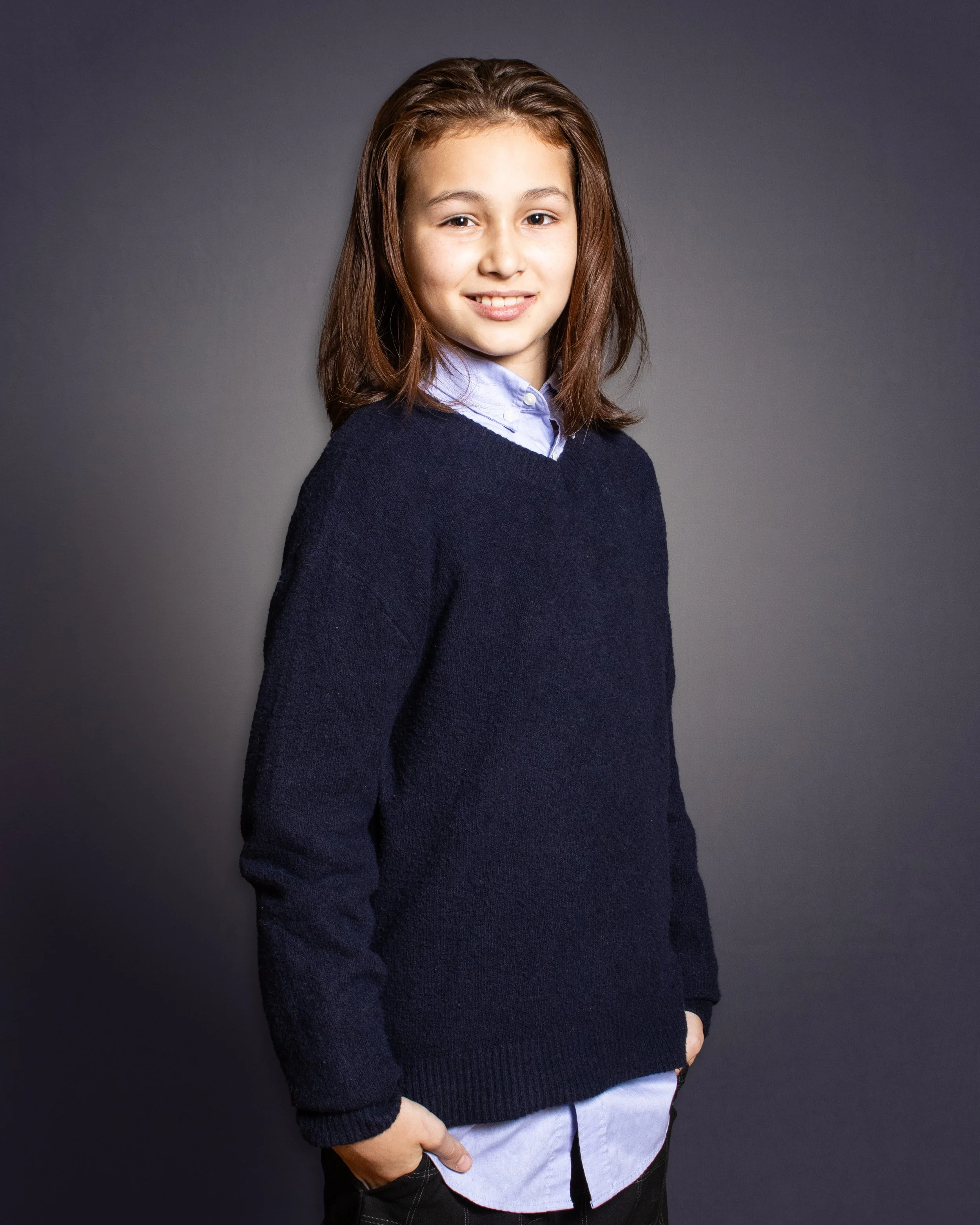 A young girl with brown hair wearing a navy sweater over a light blue collared shirt, smiling at the camera.