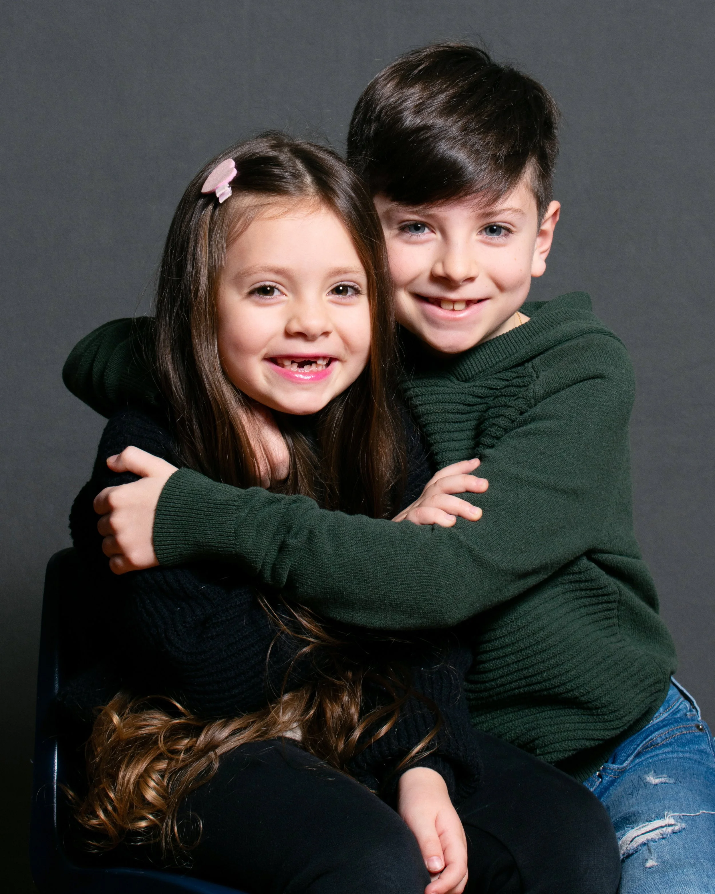 A young girl and a boy hugging and smiling at the camera against a dark grey background.