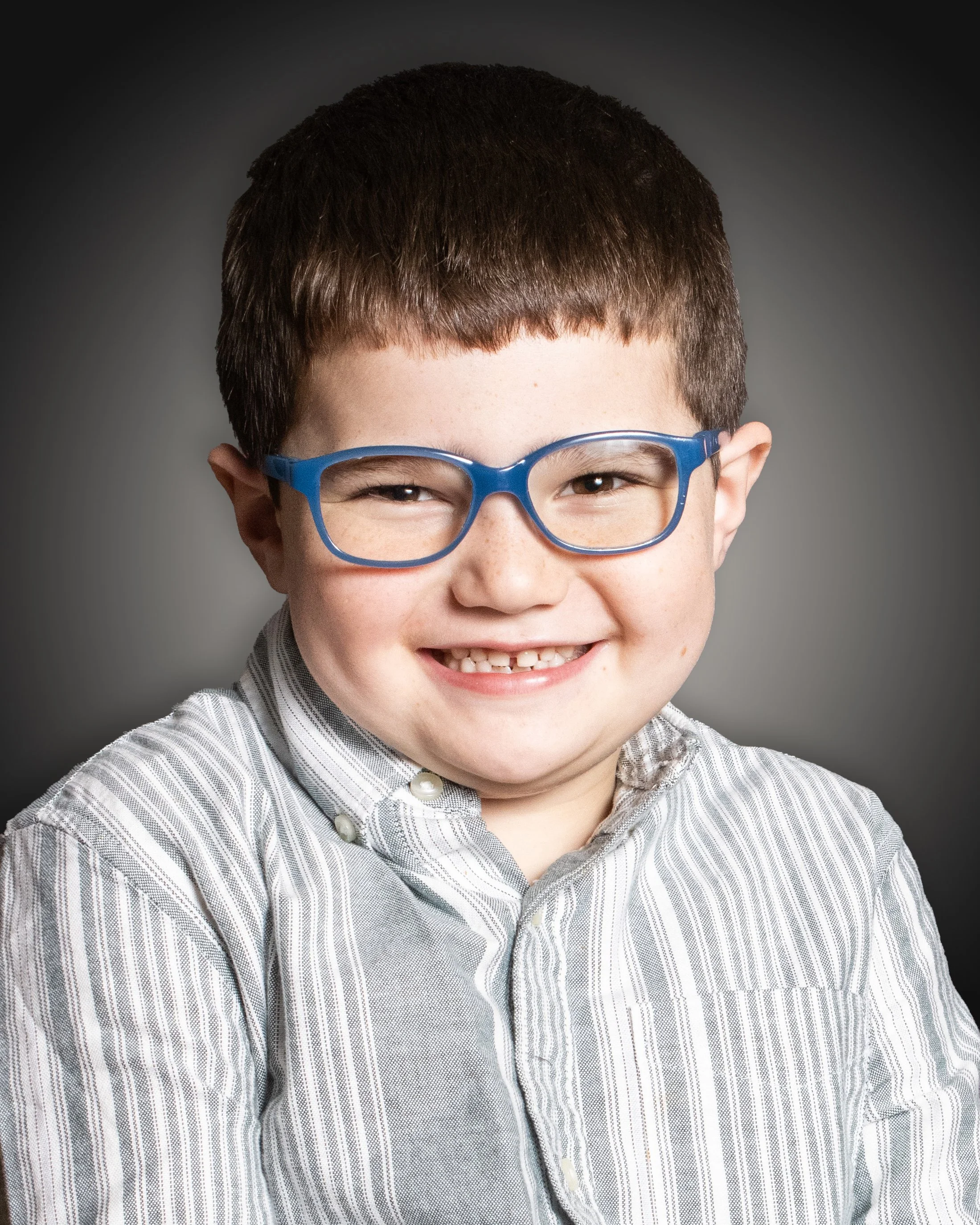 A young boy with brown hair and glasses smiling, wearing a striped shirt against a gray background.