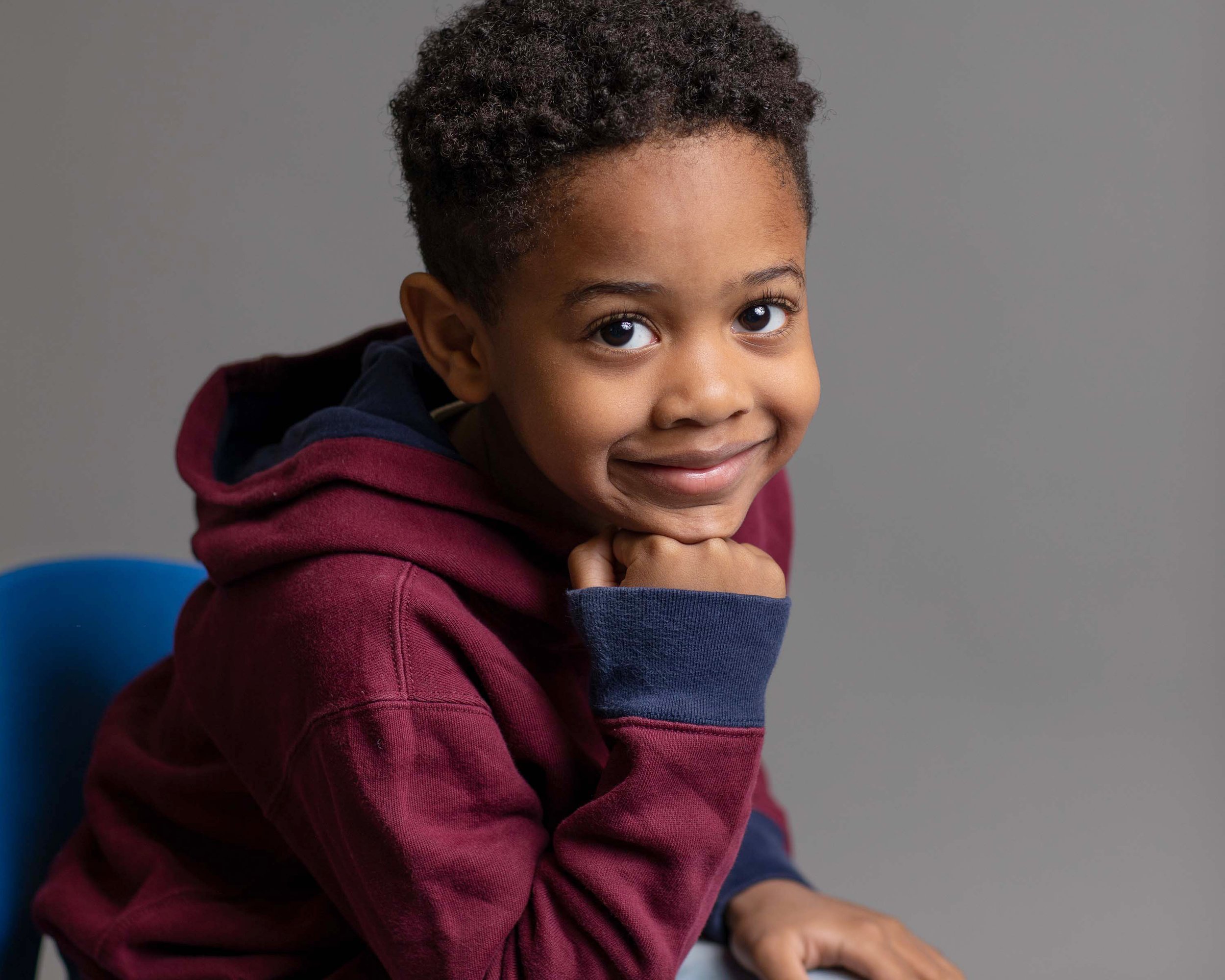 A young boy with curly hair smiling, resting his chin on his hand, wearing a maroon hoodie with navy blue cuffs, sitting on a blue chair against a plain gray background.