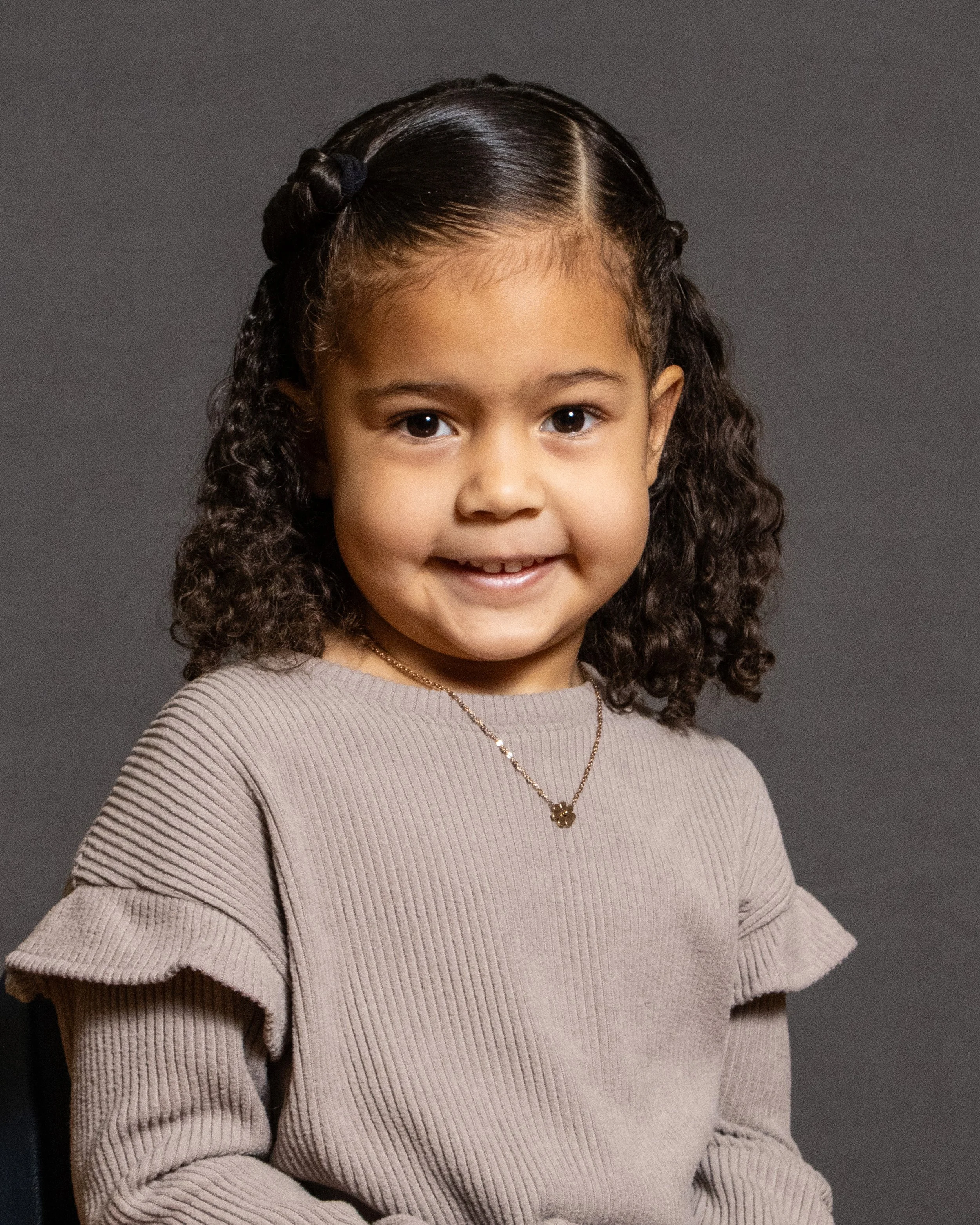 A young girl with curly dark hair, wearing a beige ruffled sweater and a necklace, smiling at the camera against a dark background.