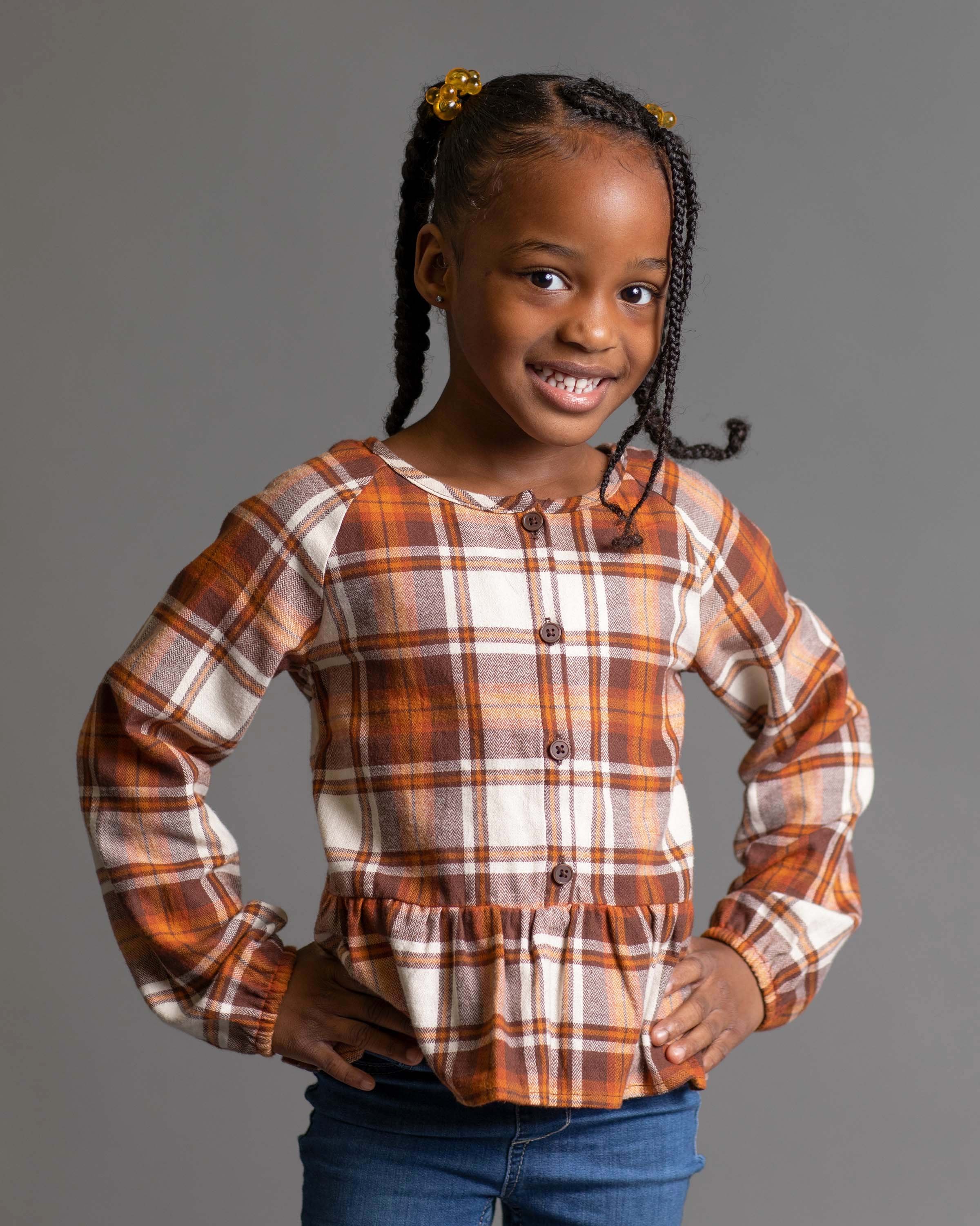 A young girl with braids decorated with yellow beads poses in front of a gray background, wearing a plaid blouse and jeans, smiling confidently with hands on hips.