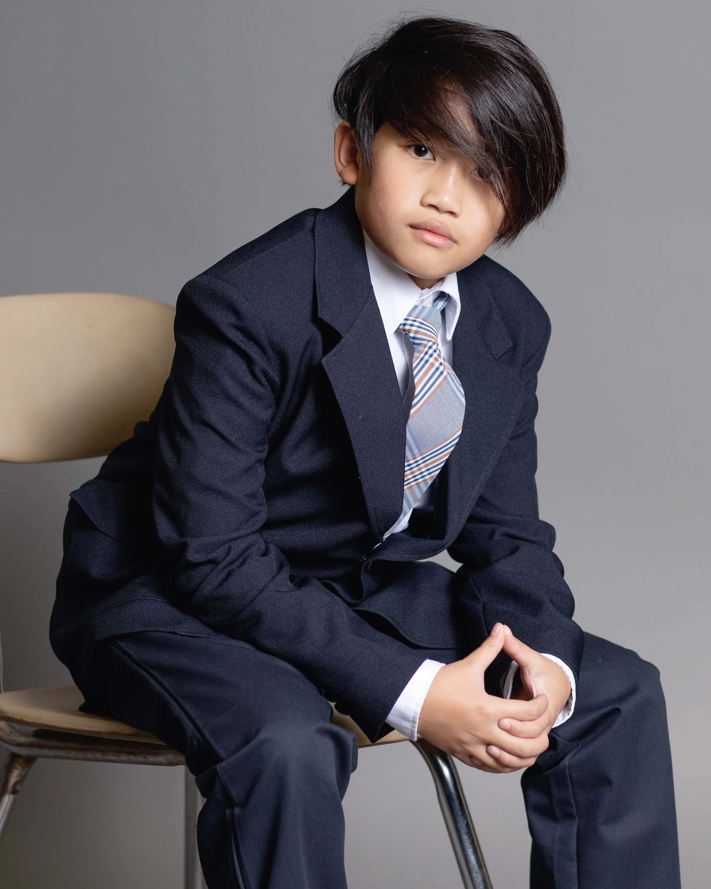 Young boy in formal business suit sitting on a beige chair against a gray background