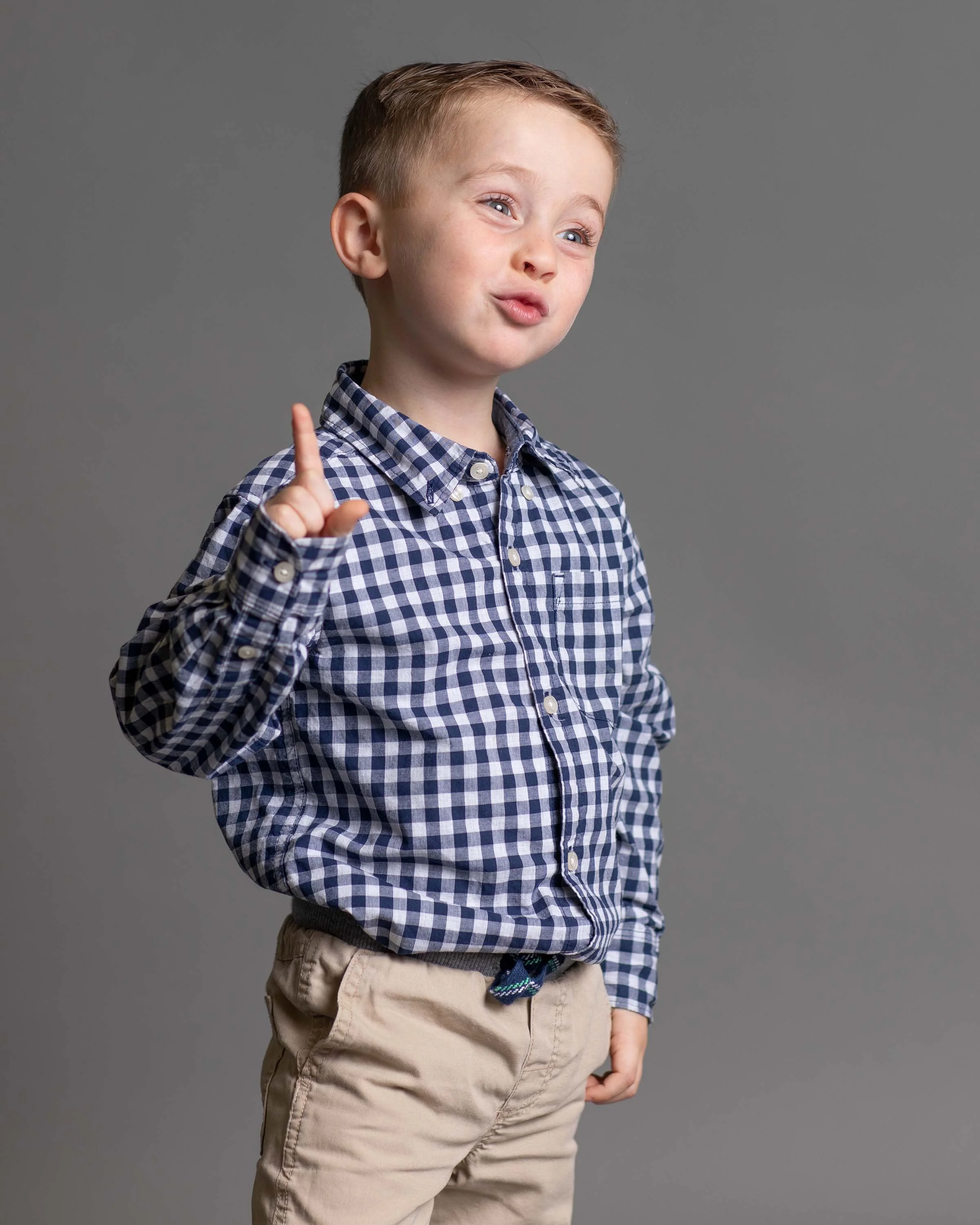A young boy with short, light brown hair wearing a blue and white checked shirt and beige pants, gesturing with his right index finger, against a plain gray background.