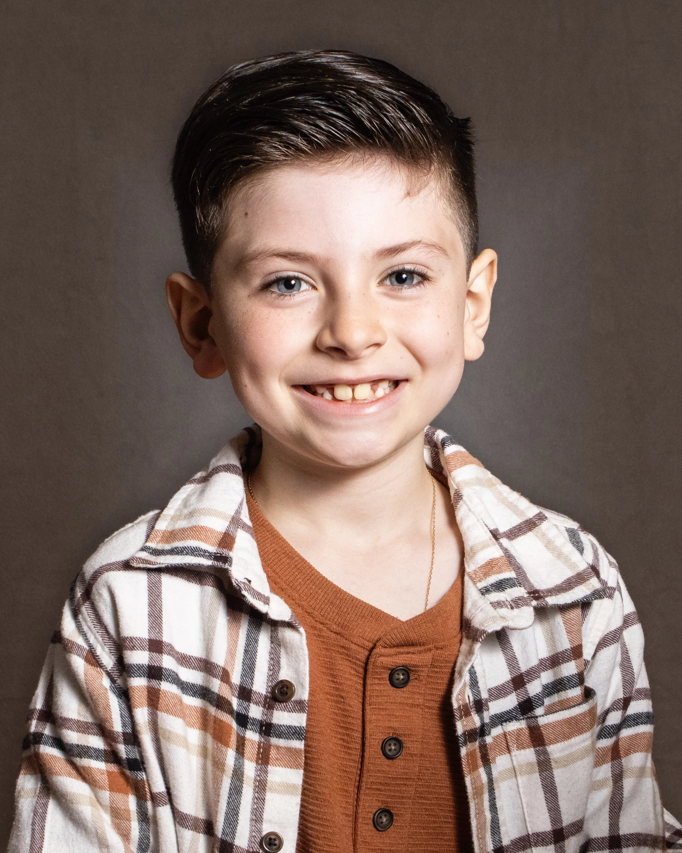 Smiling young boy with short dark hair, blue eyes, wearing a checked shirt over a rust-colored shirt, in front of a gray background.