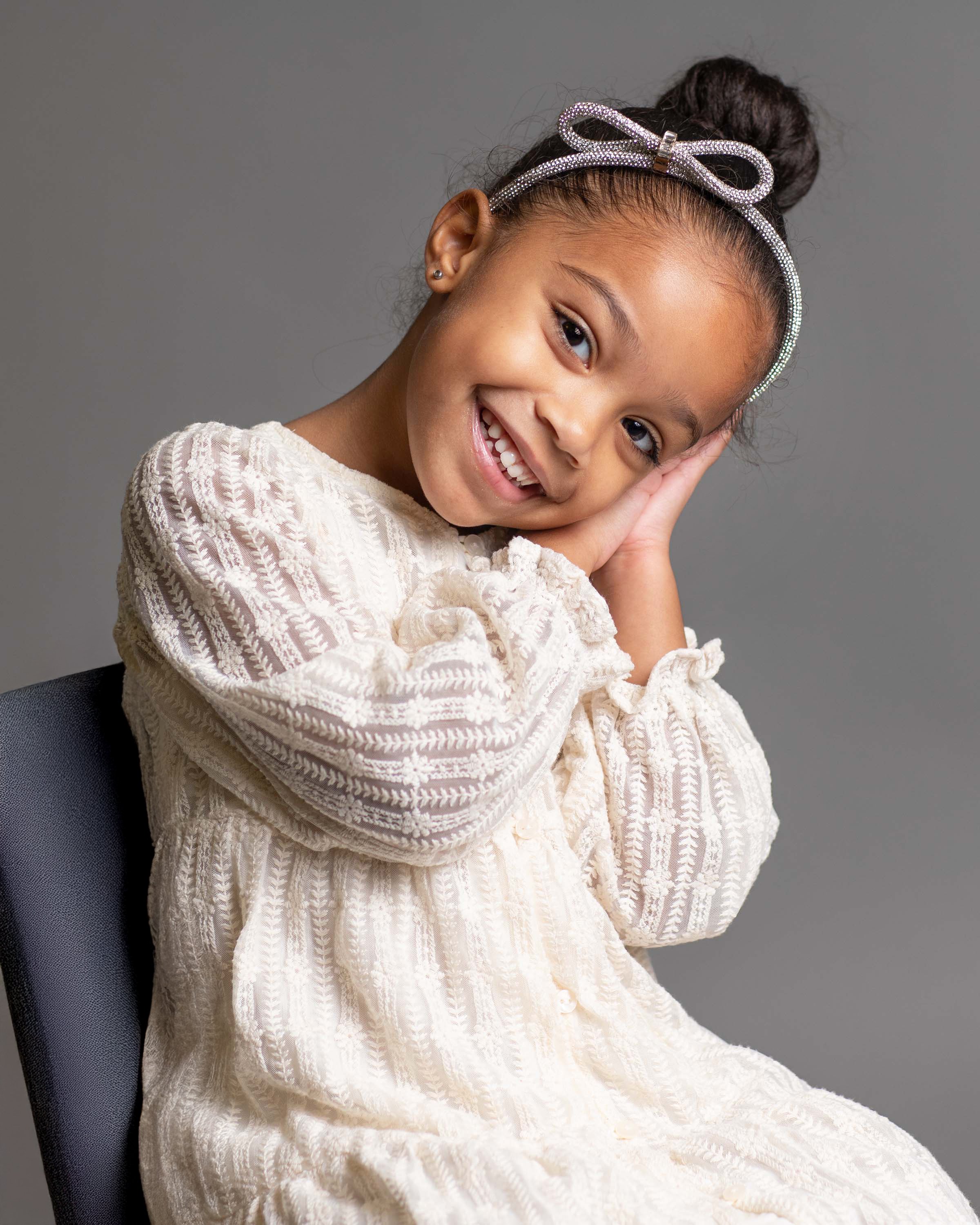 A young girl with a big smile, wearing a cream-colored dress with textured patterns, a sparkling bow headband, and having her head resting on her hand while sitting on a dark chair against a plain gray background.