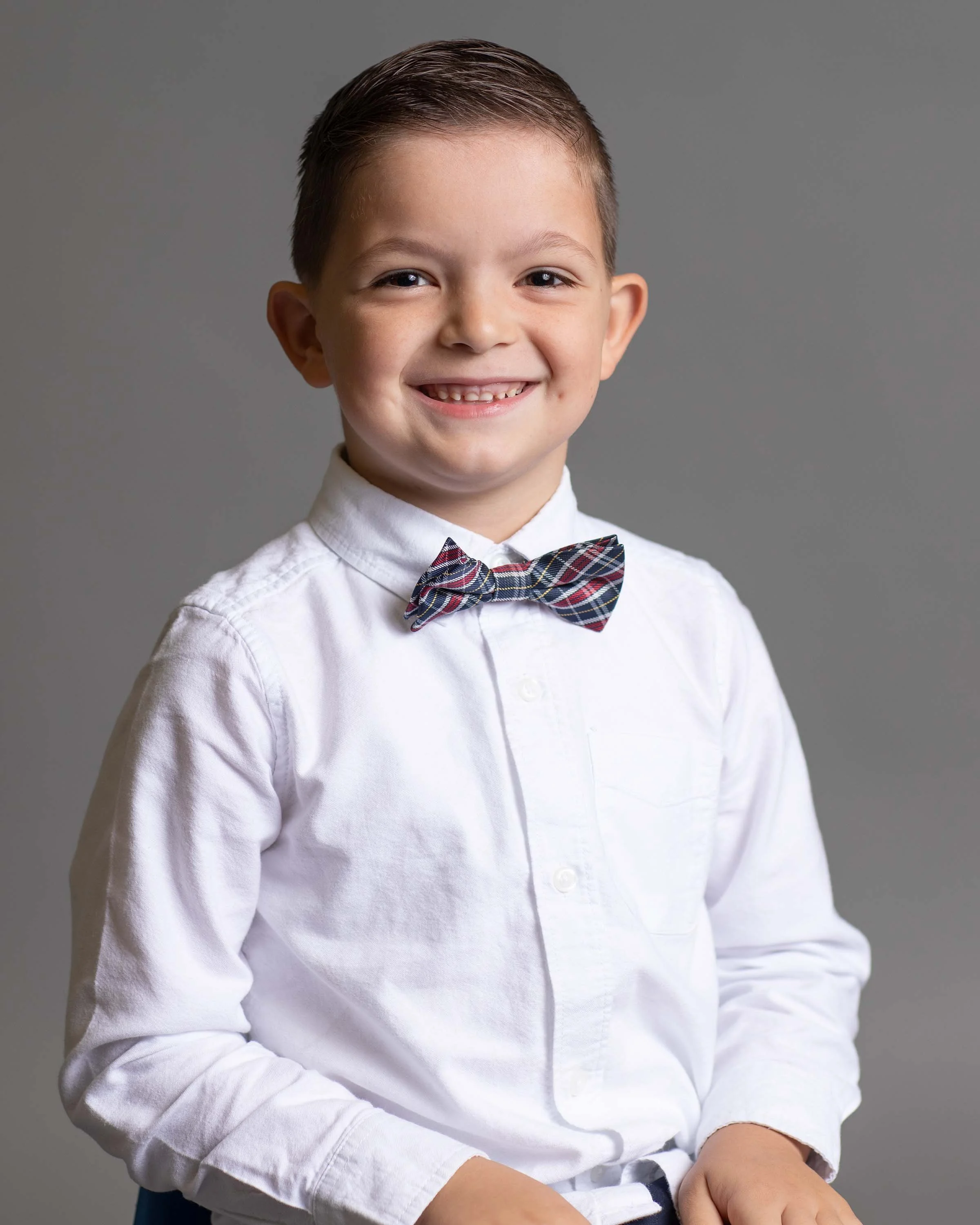 A young boy with short brown hair, wearing a white dress shirt and a plaid bow tie, smiling against a gray background.