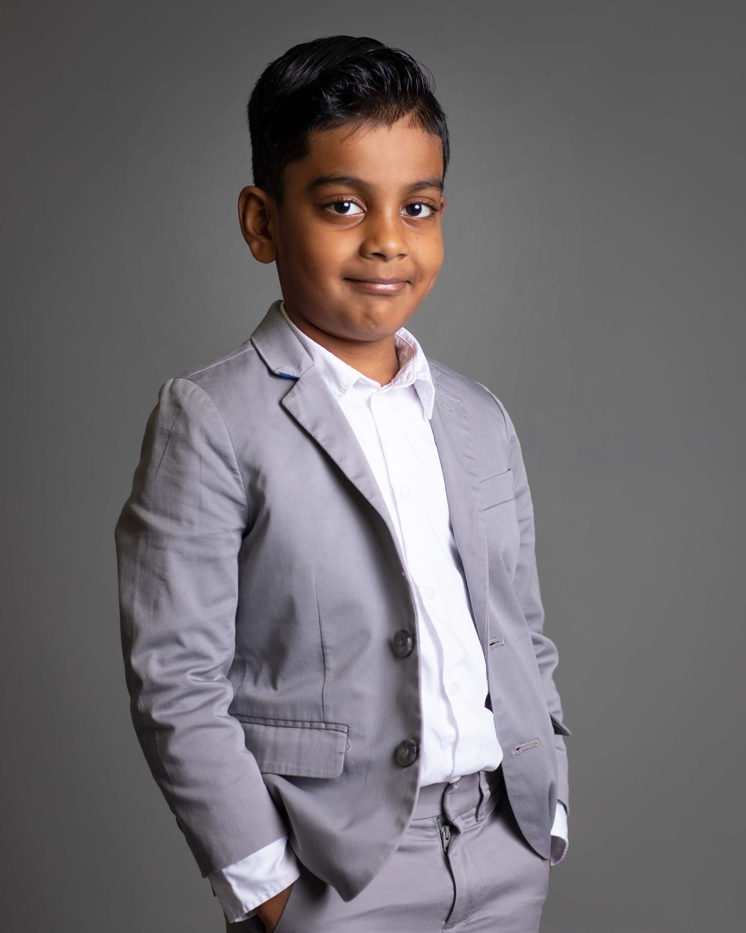 A young boy in a light gray suit with a white shirt stands against a gray background, smiling confidently.
