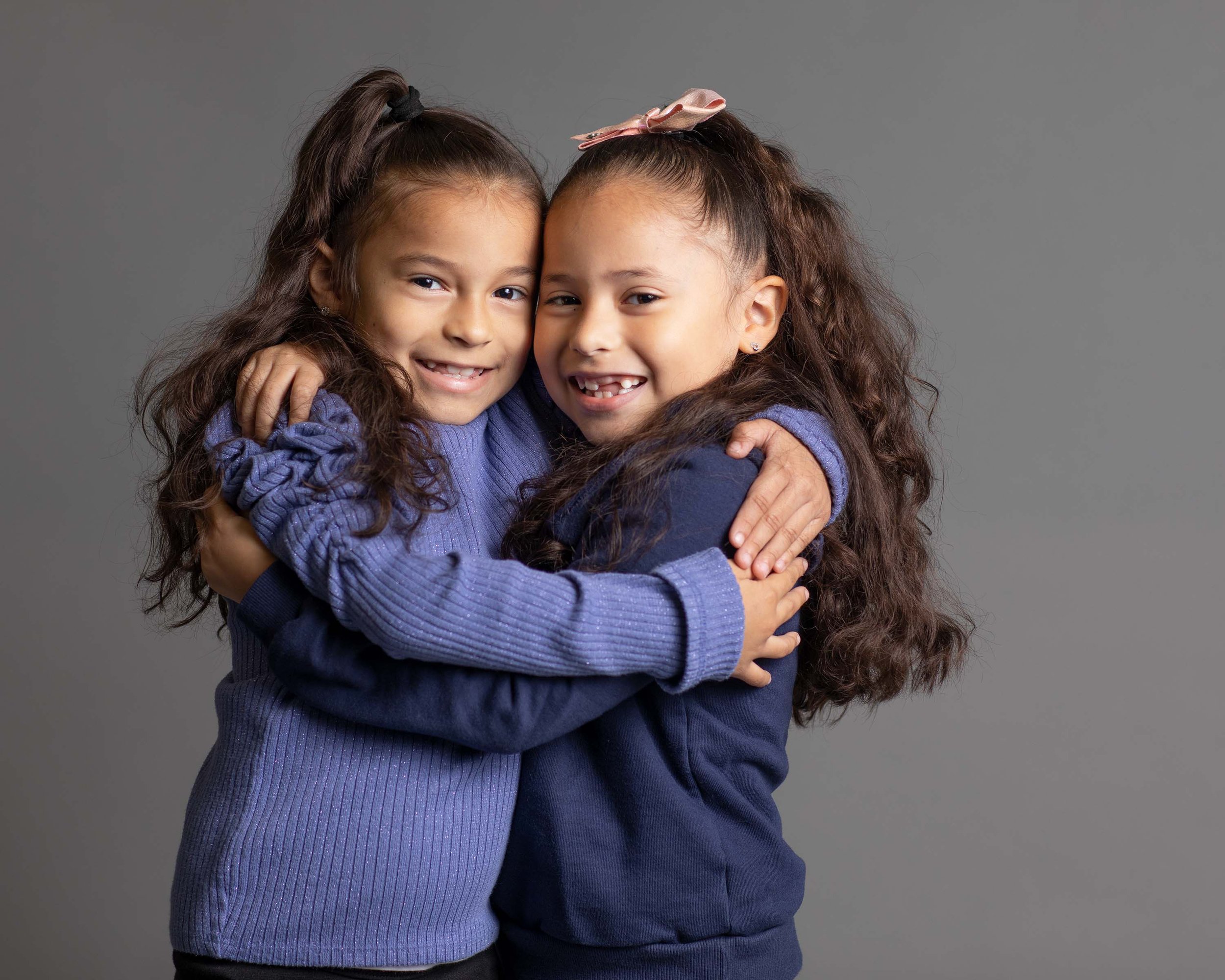 Two young girls with long curly hair hugging each other and smiling at the camera.