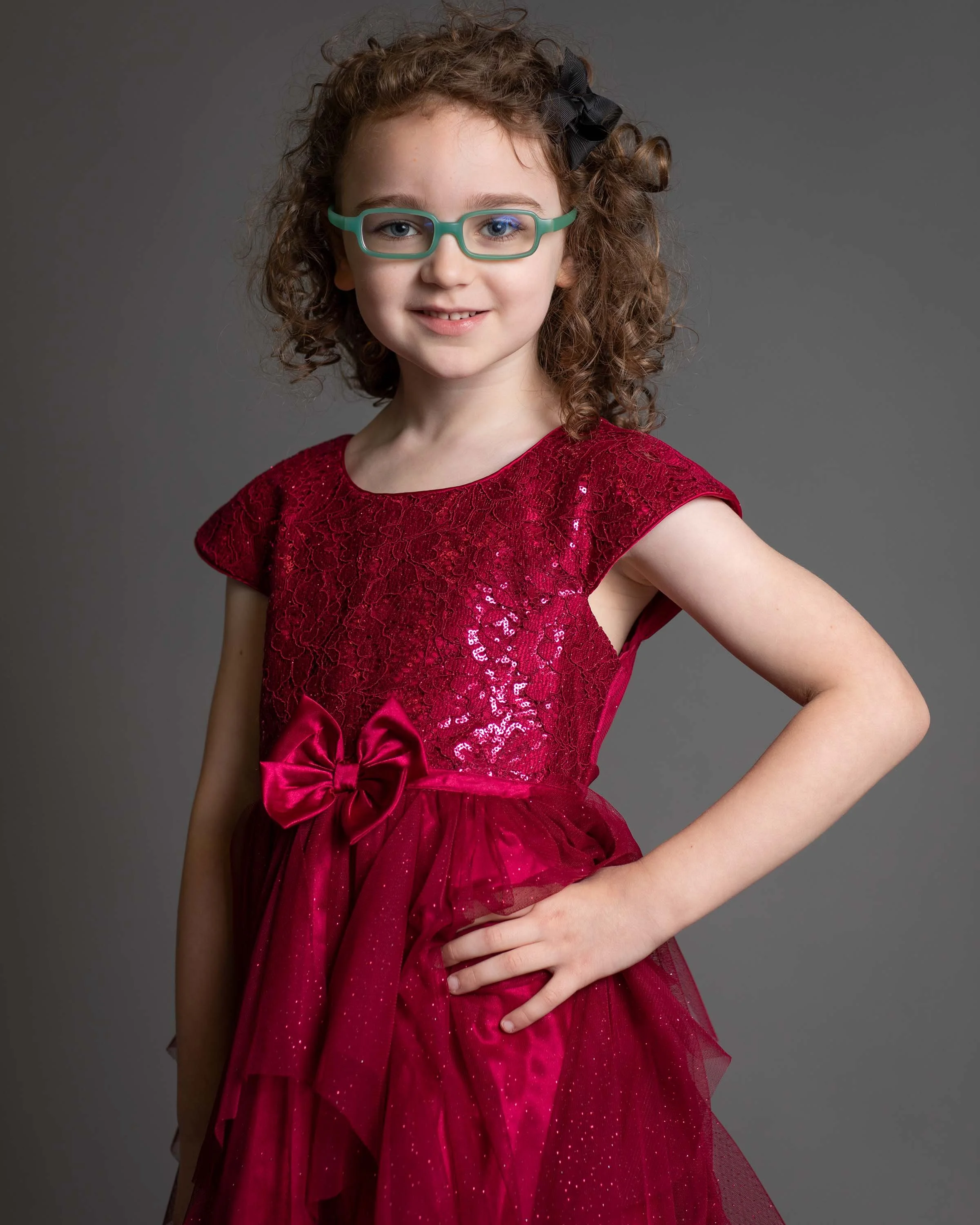 A young girl with curly hair, wearing glasses and a red dress with sequins, standing with one hand on her hip against a plain gray background.