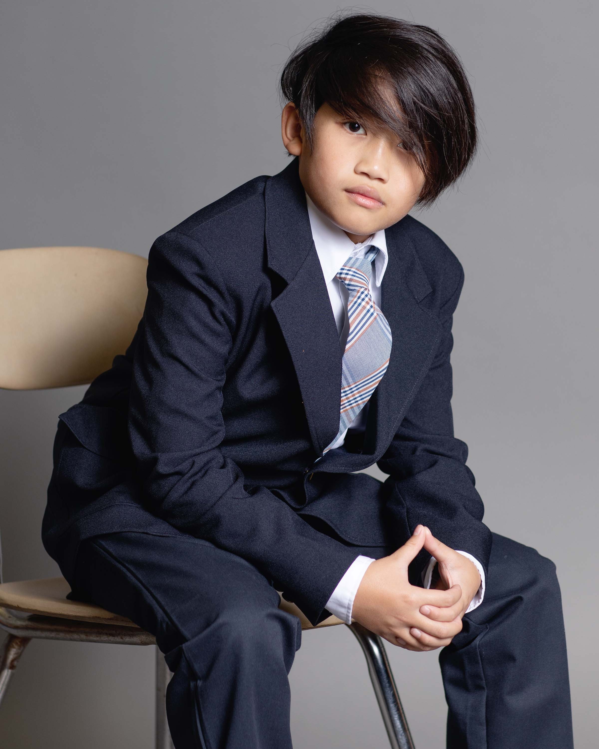 A young boy in a navy suit, white shirt, and striped tie sitting on a chair against a plain gray background.