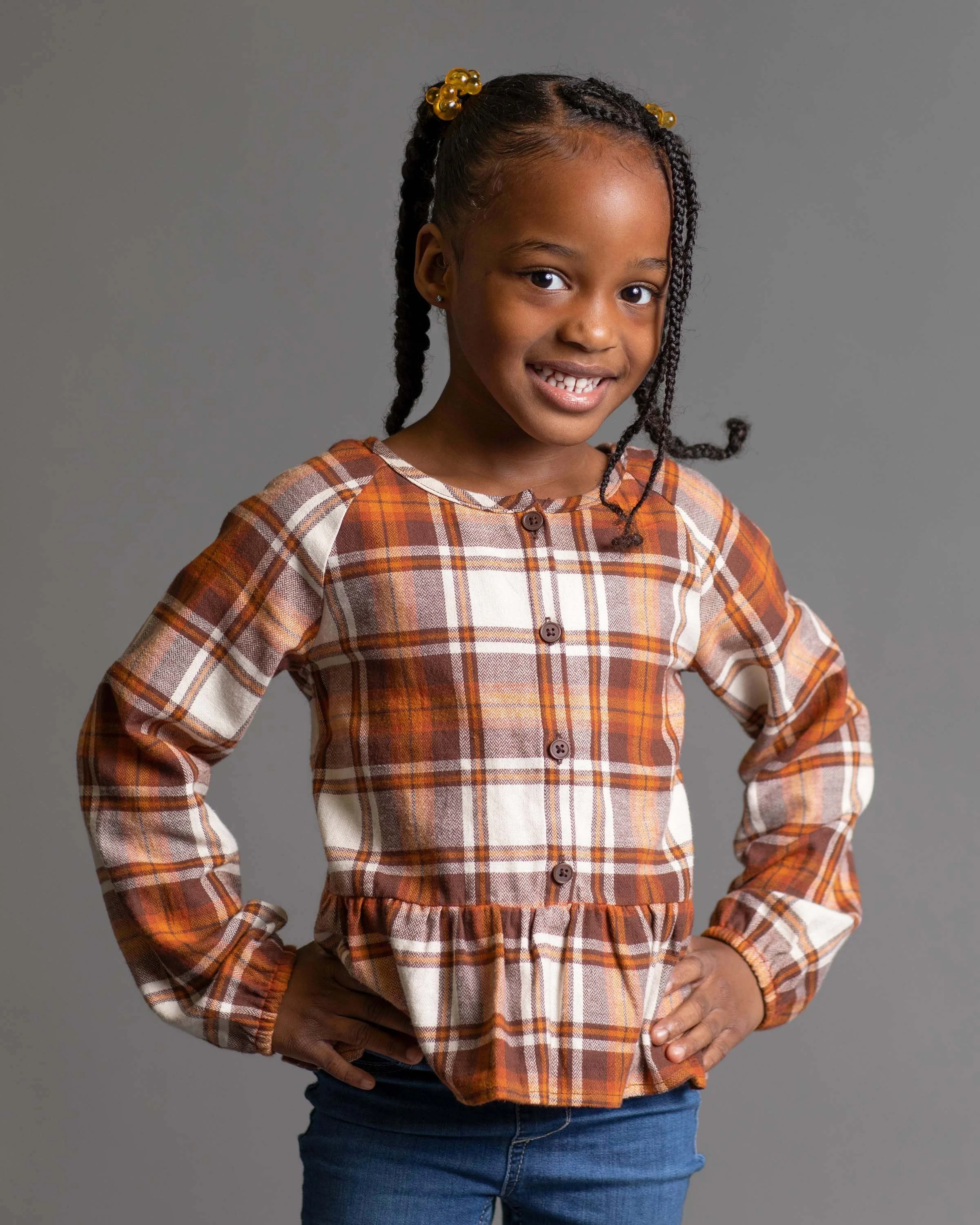A young girl with braids and yellow hair accessories, wearing a plaid shirt and blue jeans, standing with her hands on her hips and smiling.
