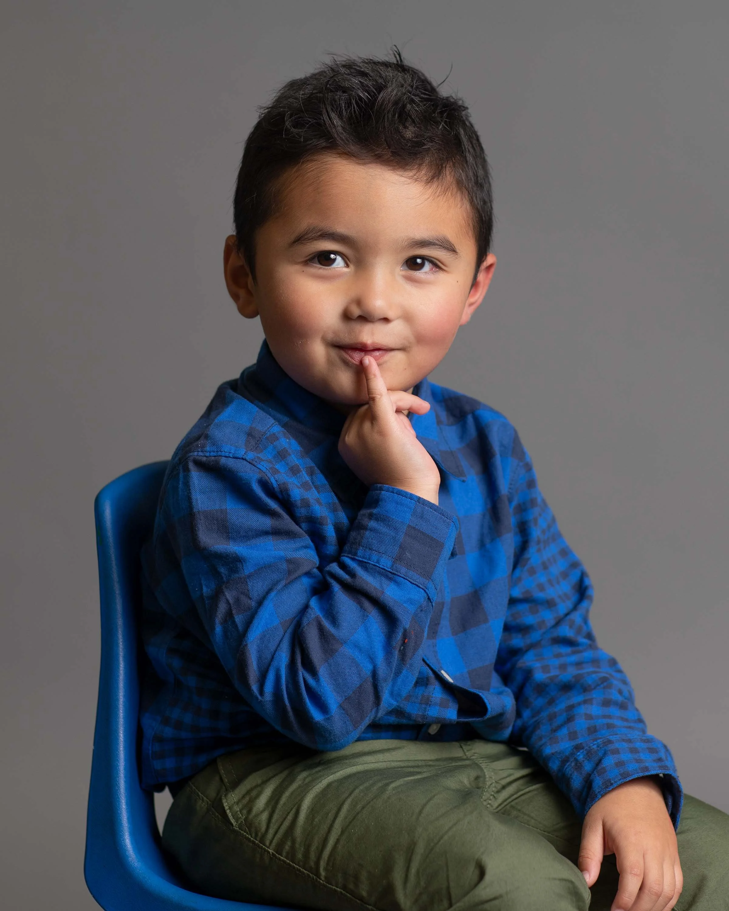 A young boy with short dark hair, wearing a blue plaid shirt and olive green pants, sitting on a blue chair against a gray background. He has a playful expression, with one finger on his lips, as if thinking or posing.