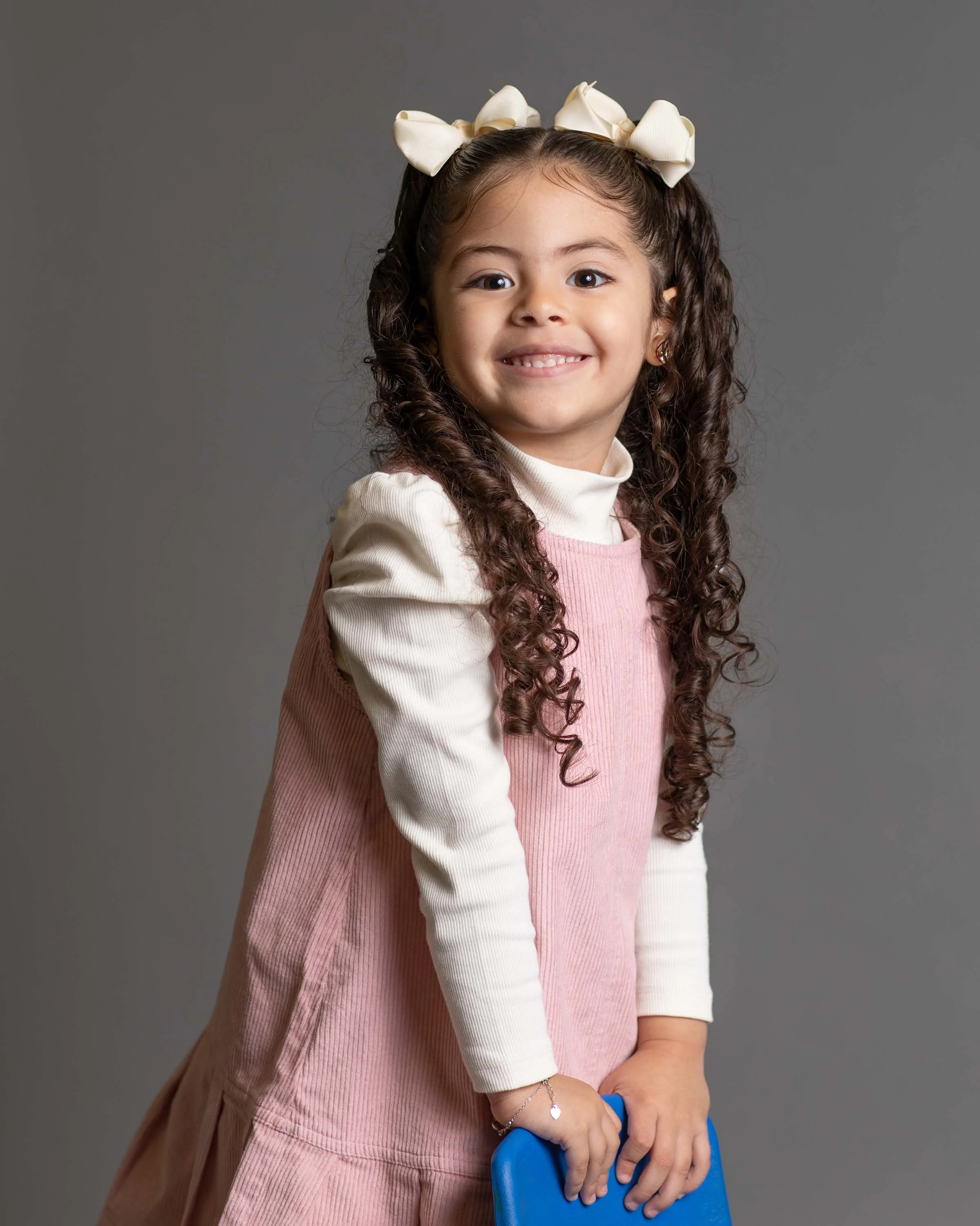 A young girl with curly brown hair, wearing a white bow on her head, a white long sleeve shirt under a pink sleeveless dress, smiling and holding a blue book against a plain grey background.