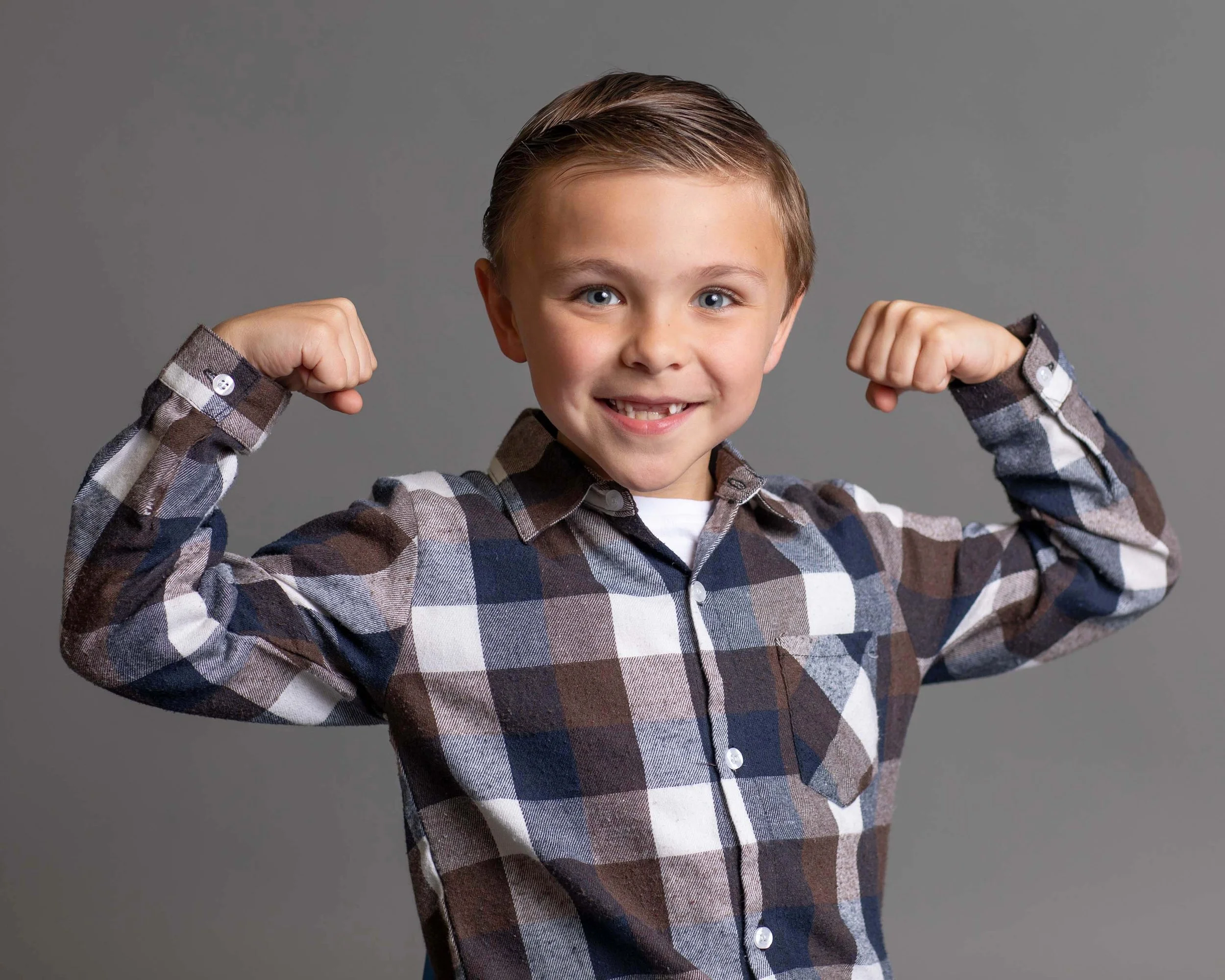 A young boy with blond hair, blue eyes, and a big smile, flexing his arm muscles, wearing a brown, white, and blue plaid shirt, against a gray background.