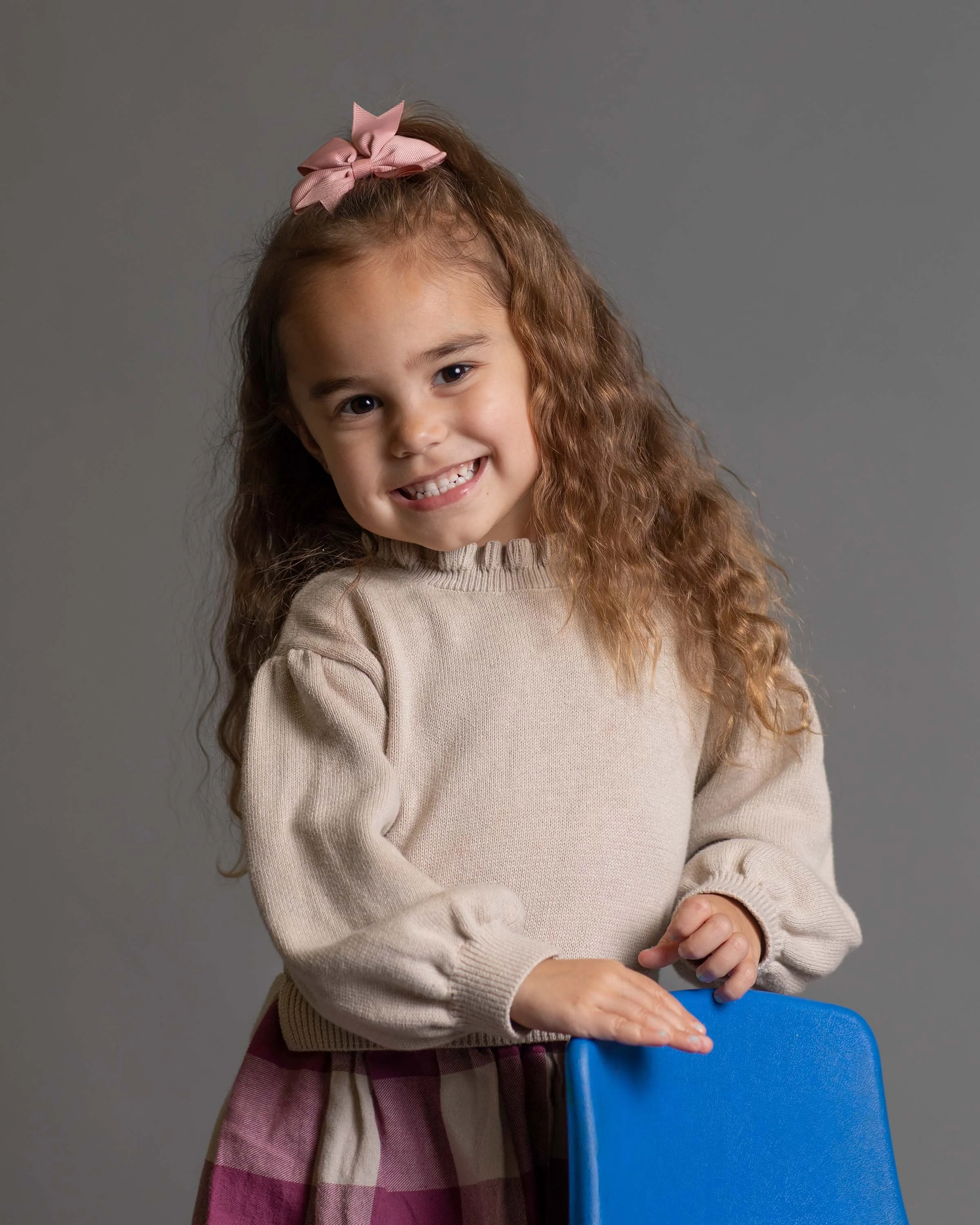Young girl with curly hair, wearing a pink bow, beige sweater, and a pink plaid skirt, smiling and holding onto a blue chair against a gray background.