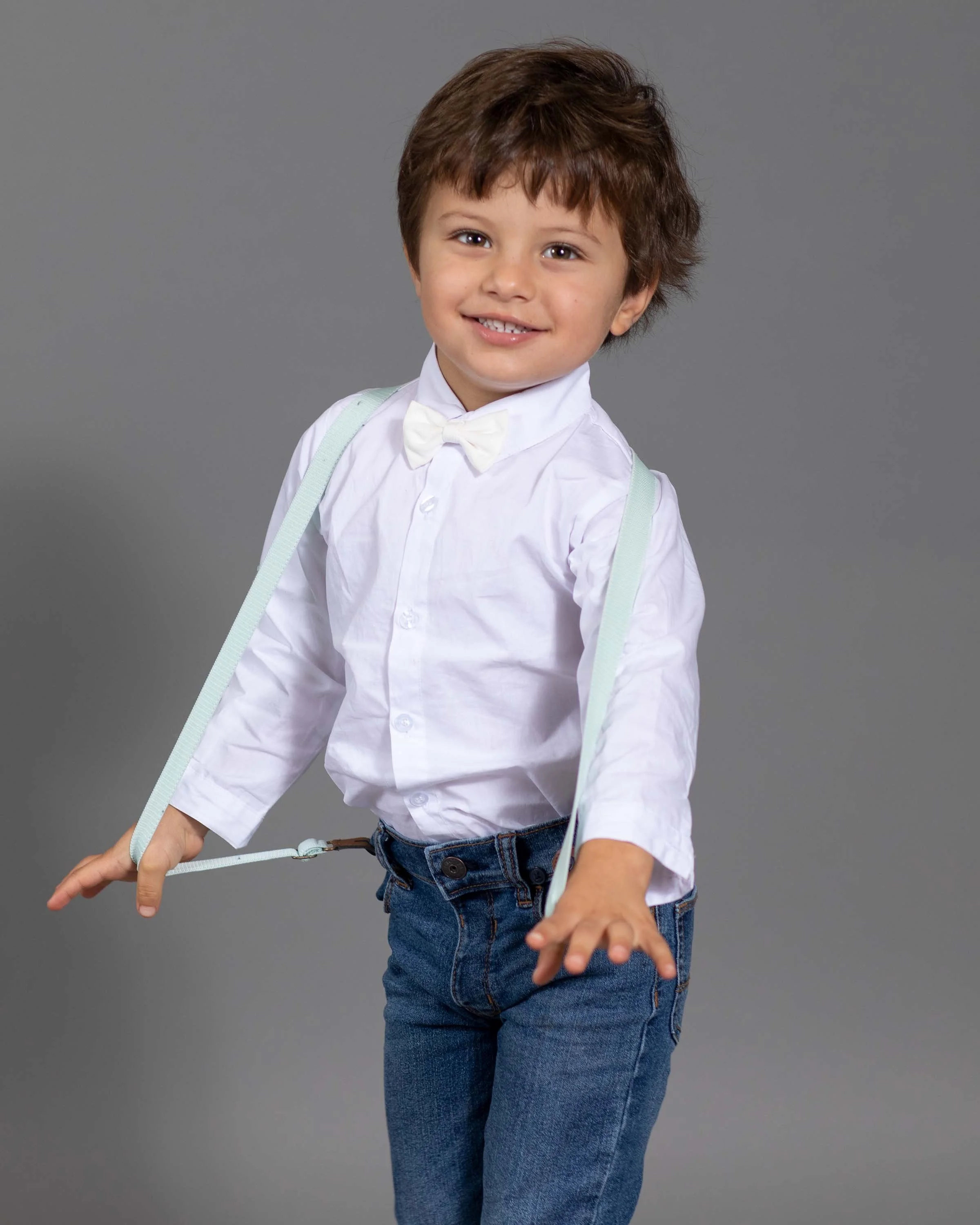 Young boy with brown hair smiling, dressed in white shirt, bow tie, jeans, and suspenders, posing against a gray background