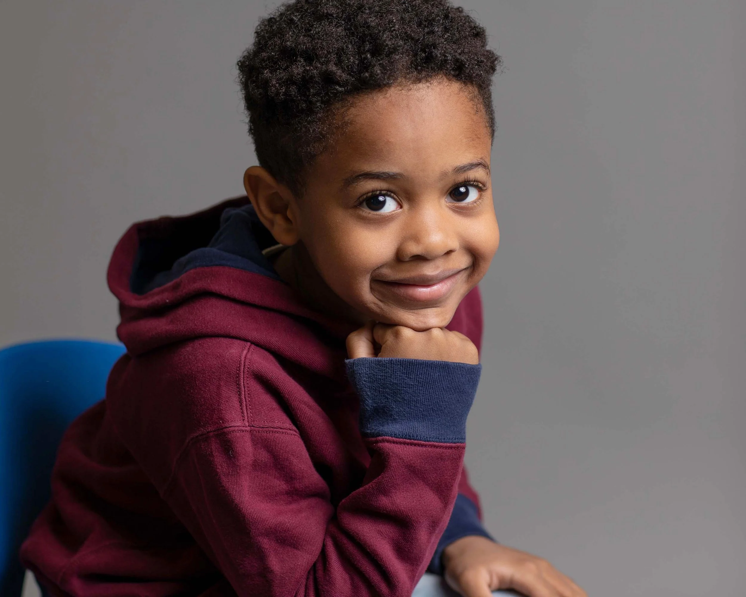 A young boy with curly hair smiling, wearing a maroon hoodie and sitting on a blue chair against a plain gray background.