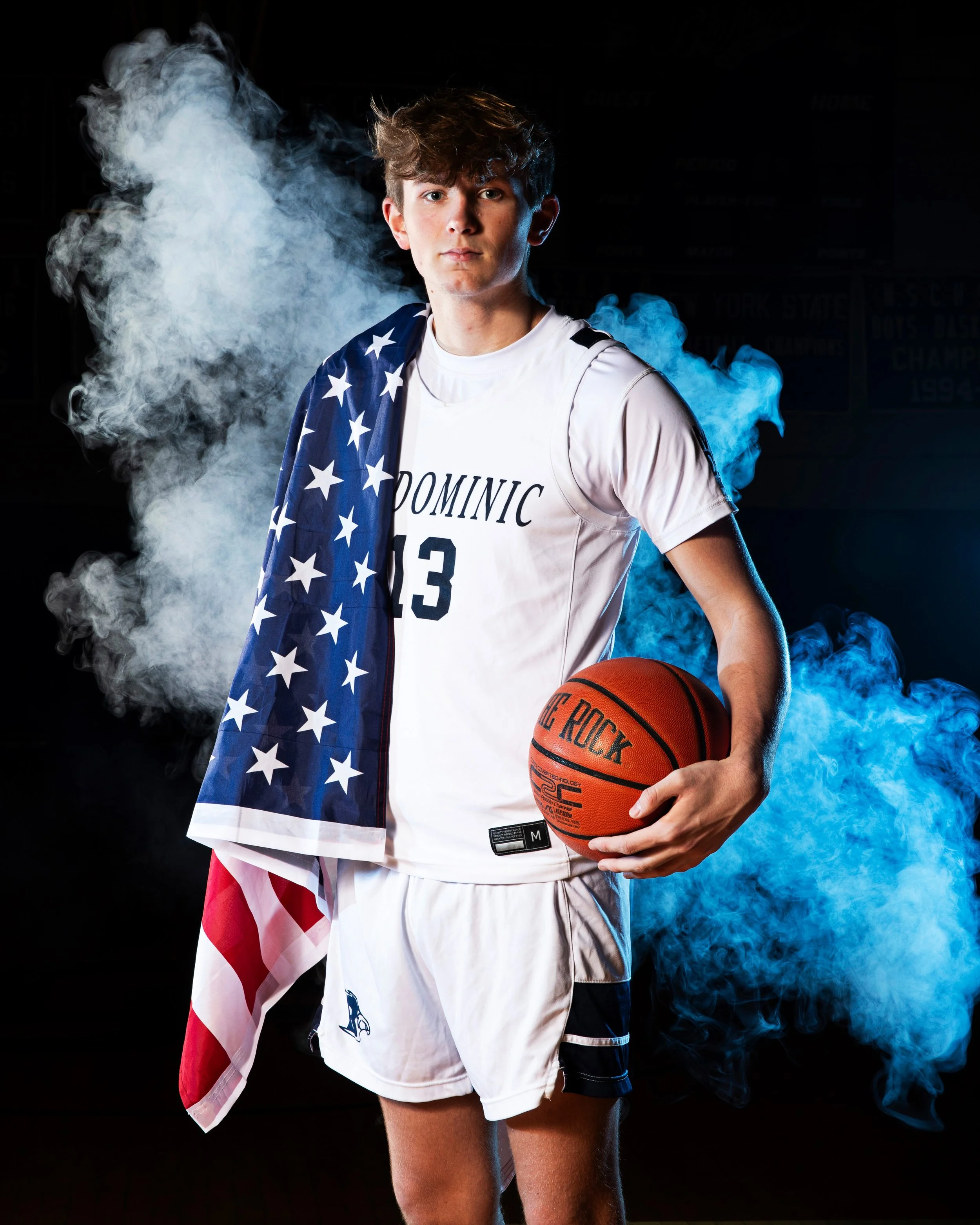 Young male basketball player holding a basketball, draped with an American flag, wearing a jersey with 'Dominic 13,' standing in front of a smoky background.