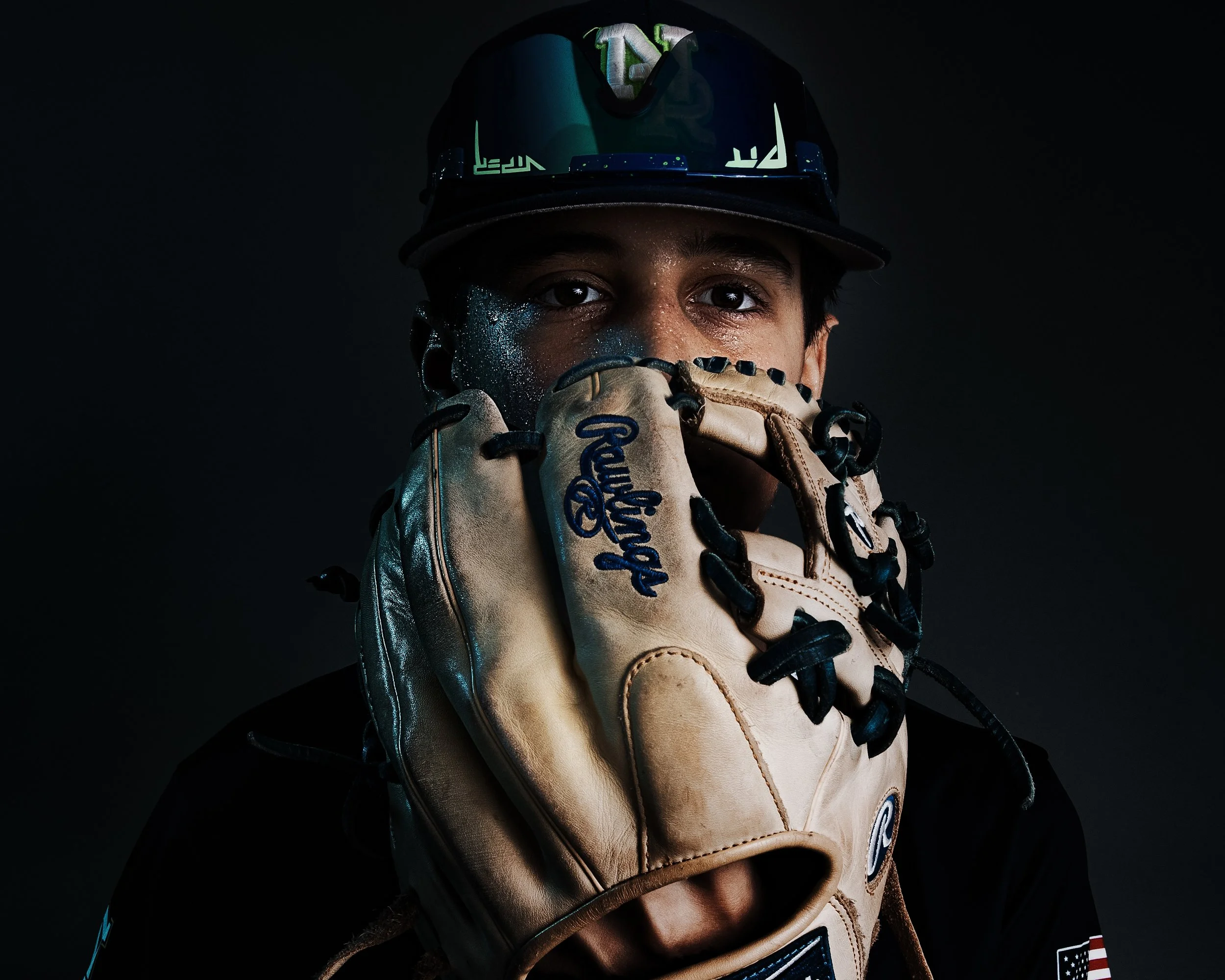 A baseball player wearing a black helmet with green accents and a Rawlings glove, covering part of his face with his gloved hand and looking directly at the camera against a dark background.