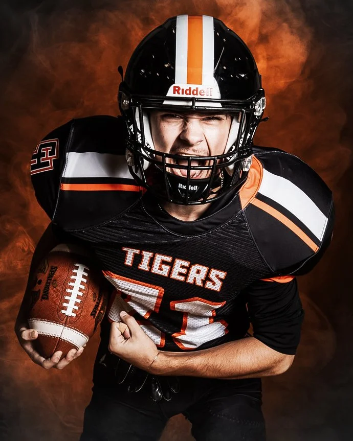 An American football player in black uniform with orange and white accents, wearing a helmet, holding a football, and making an intense facial expression with a smoky orange background.