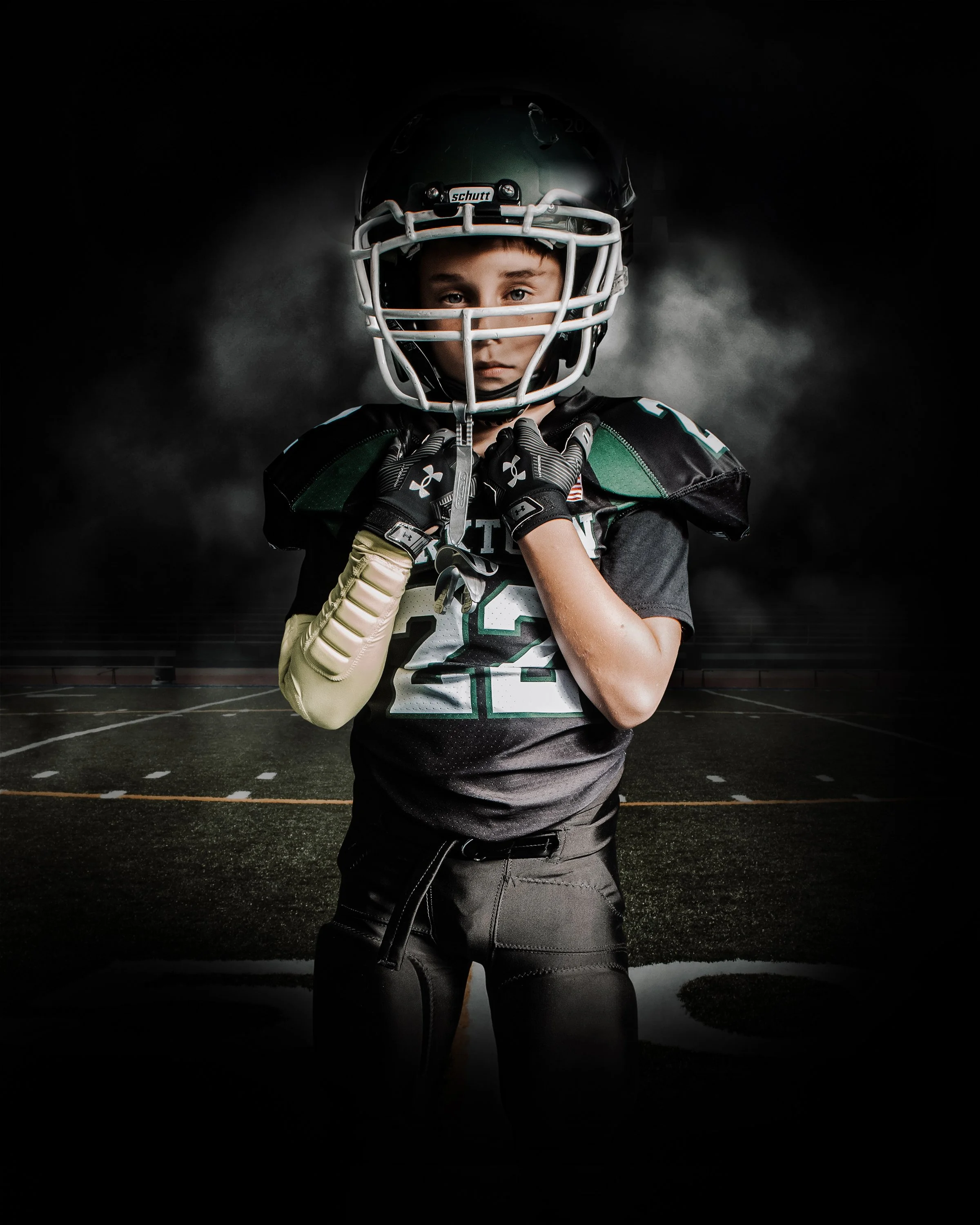 Young boy in football uniform and helmet standing on a football field at night, looking serious.