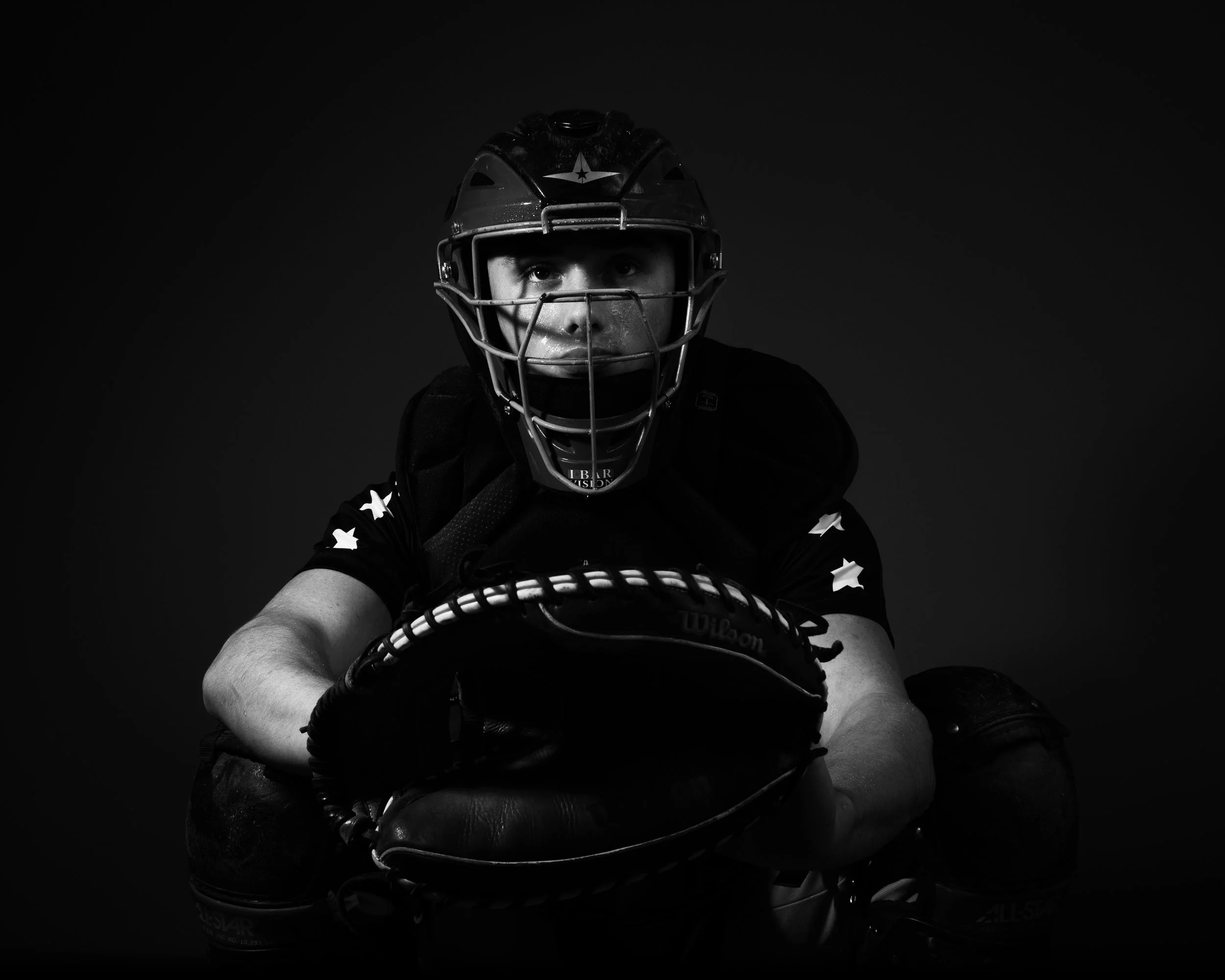 Black and white photo of a male baseball catcher in gear, crouching with glove, wearing a helmet and protective face mask, against a dark background.