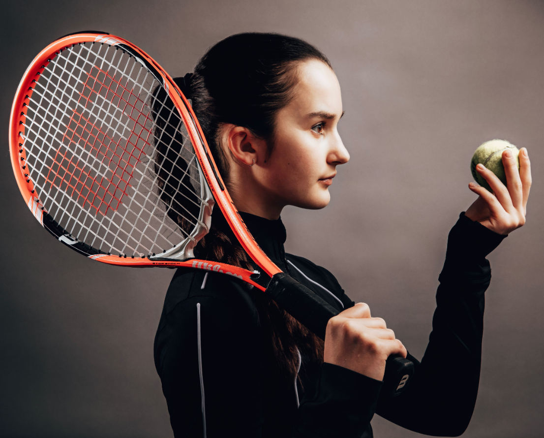 A young woman with dark hair tied back, holding a tennis racket over her shoulder with her right hand, and a tennis ball in her left hand, looking at the ball with a focused expression.